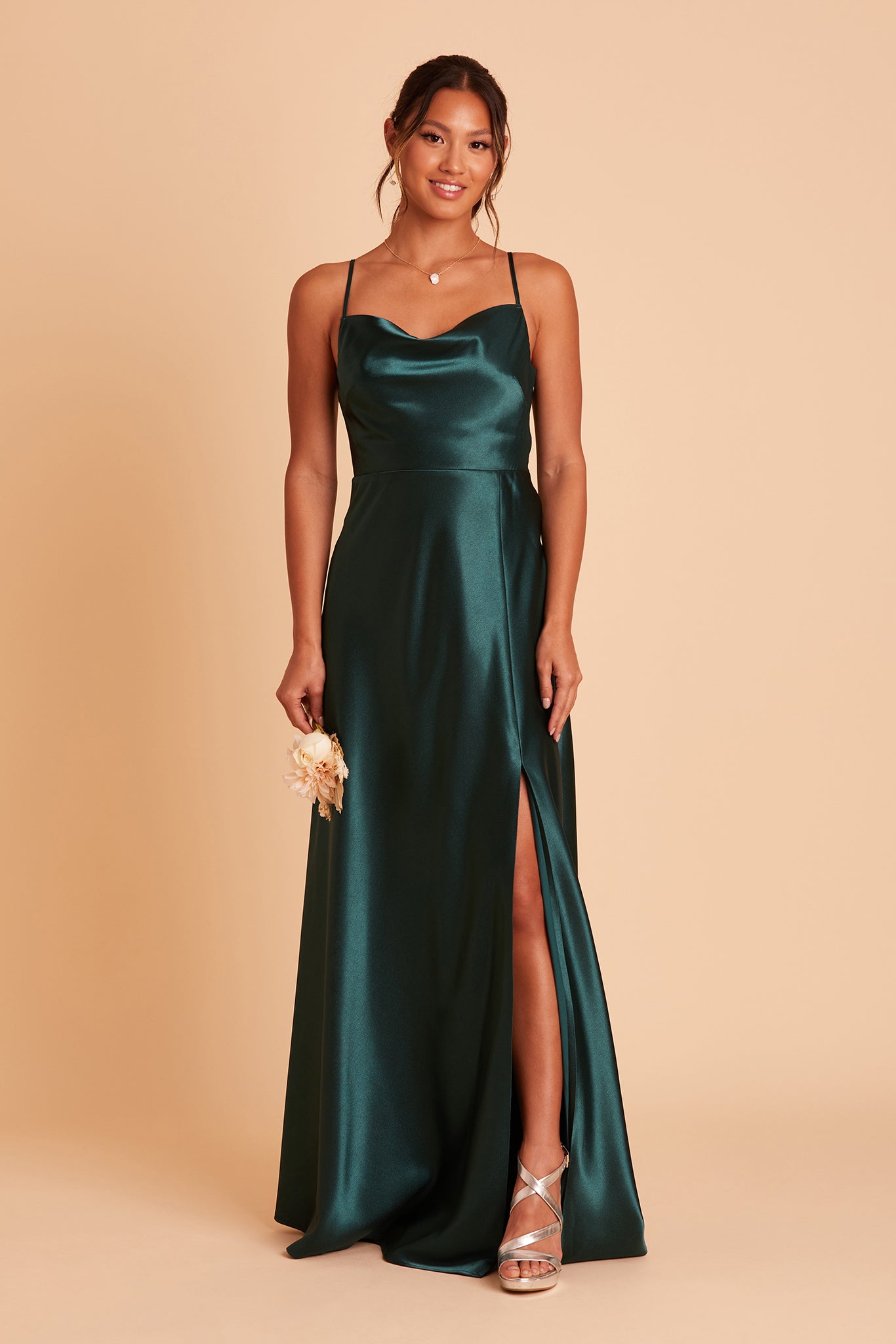 Front view of the Lisa Long Dress in emerald satin shows a slender model with a medium skin tone wearing a lightly draped cowl neck bodice with spaghetti straps and a floor-length flared dress with a slit. 