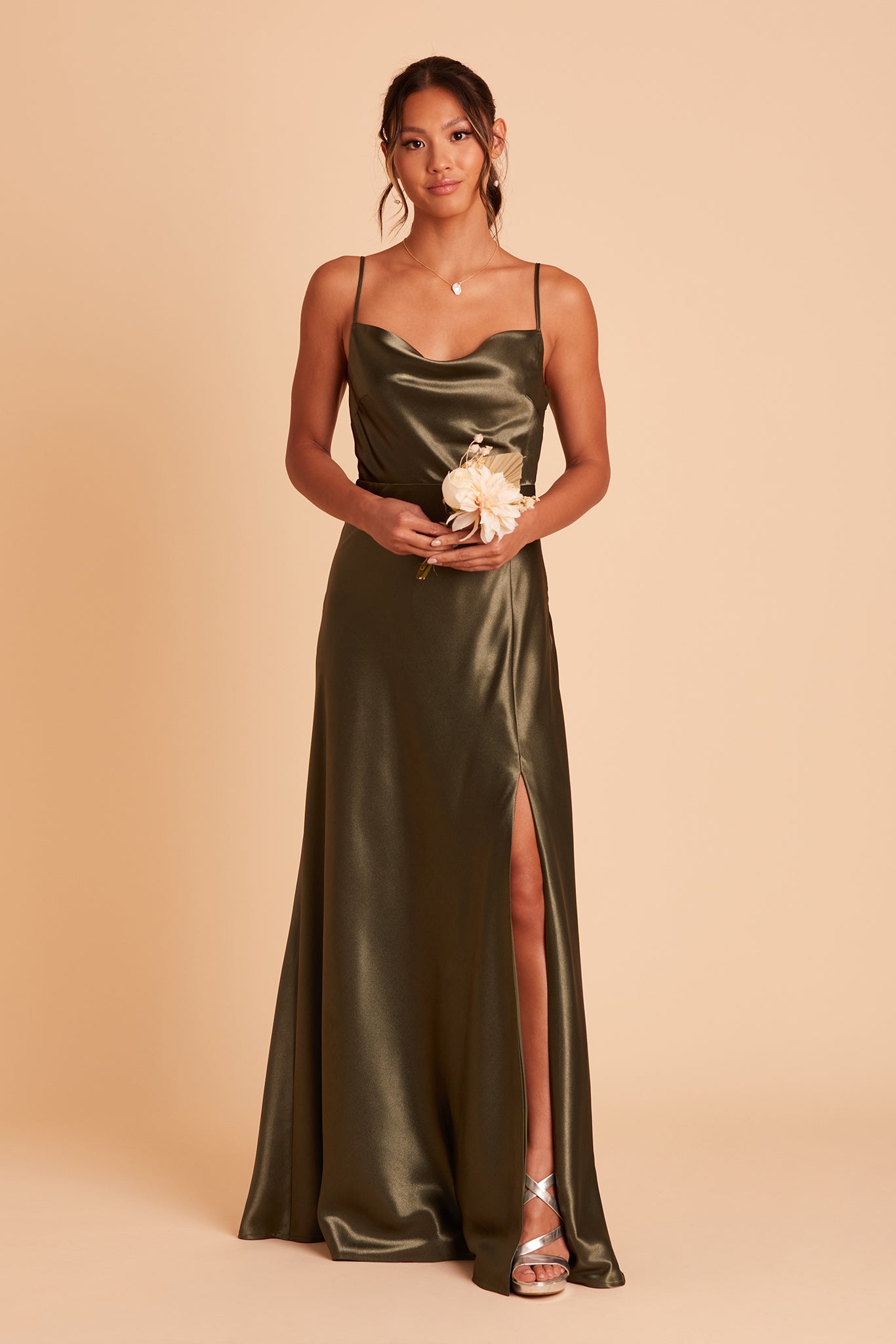 Front view of the Lisa Long Dress in olive satin shows a model revealing their leg and foot in the mid-thigh slit. They wear strappy high heel shoes in platinum metallic.