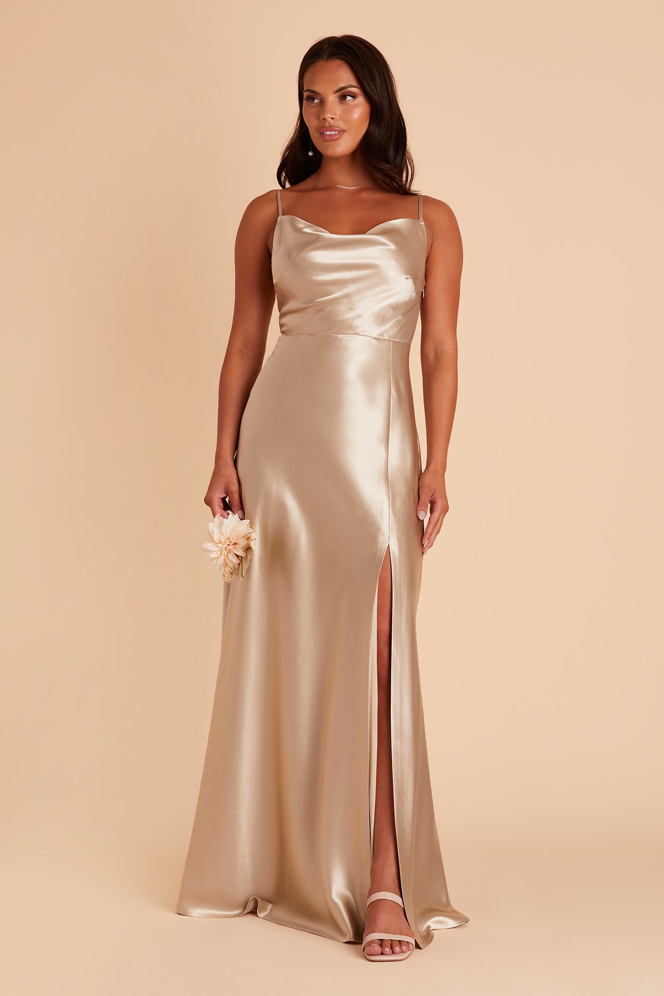 Front view of the Lisa Long Dress in neutral champagne satin shows a slender model with a medium skin tone wearing a lightly draped cowl neck bodice with spaghetti straps and a floor length flared dress with a slit. 