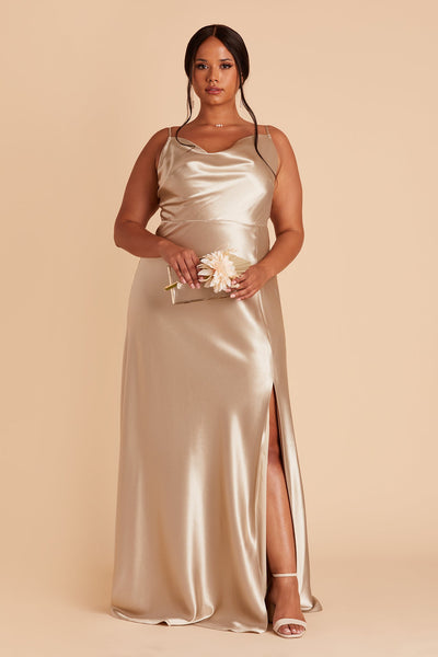 Front view of the Lisa Long Dress Curve in neutral champagne satin shows a full-figured model with a medium skin tone wearing a lightly draped cowl neck bodice with spaghetti straps and a floor length flared dress with a slit. They hold the  Clear Clutch purse.
