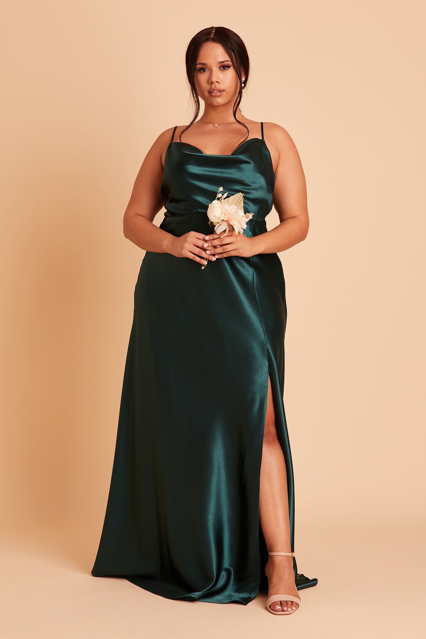 Front view of the Lisa Long Dress Curve in emerald satin shows a full-figured model with a medium skin tone wearing a lightly draped cowl neck bodice with spaghetti straps and a floor length flared dress with a slit. 