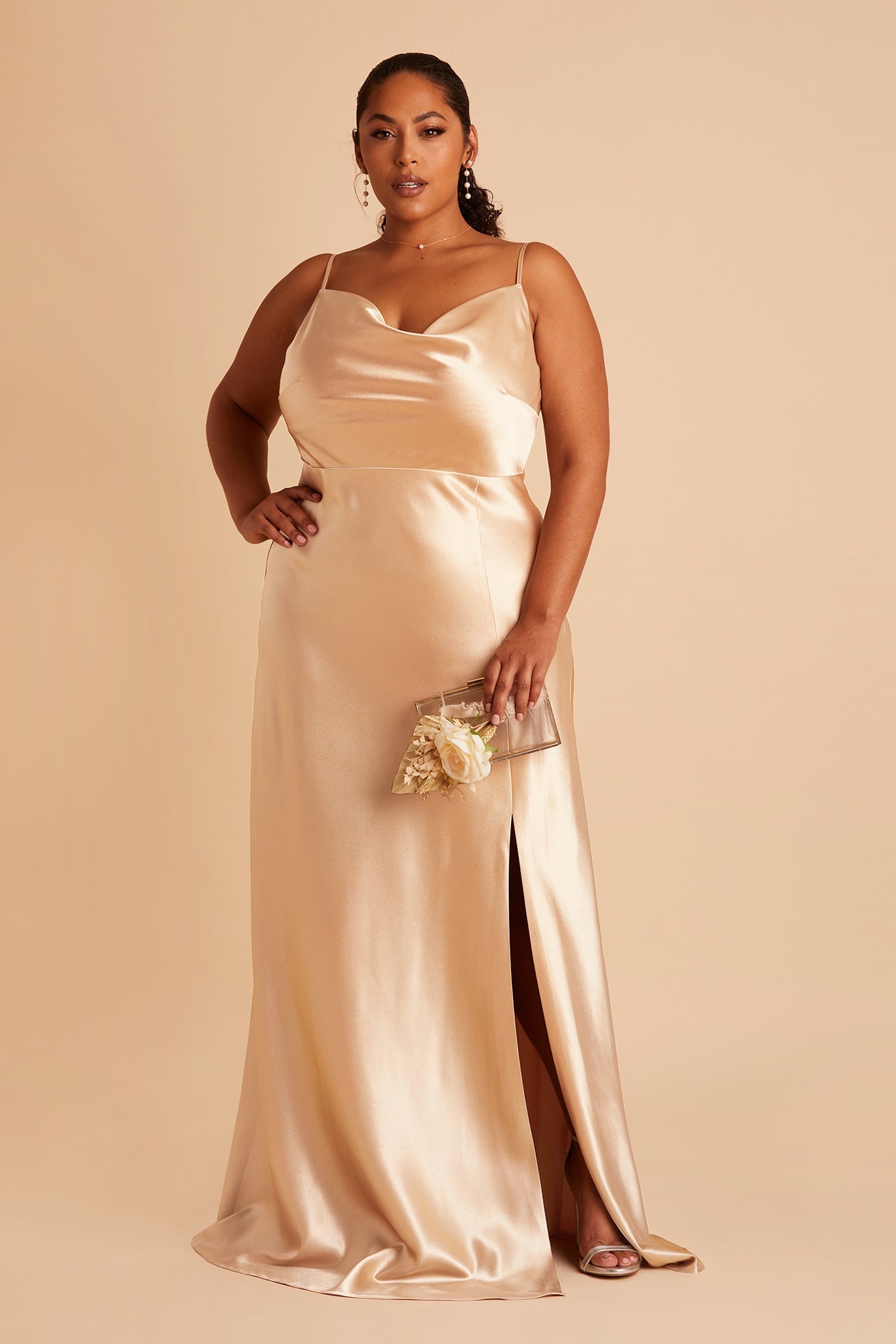 Front view of the Lisa Long Dress Curve in gold satin shows a full-figured model with a medium skin tone wearing a lightly draped cowl neck bodice with spaghetti straps and a floor length flared dress with a slit. They hold the  Clear Clutch purse.