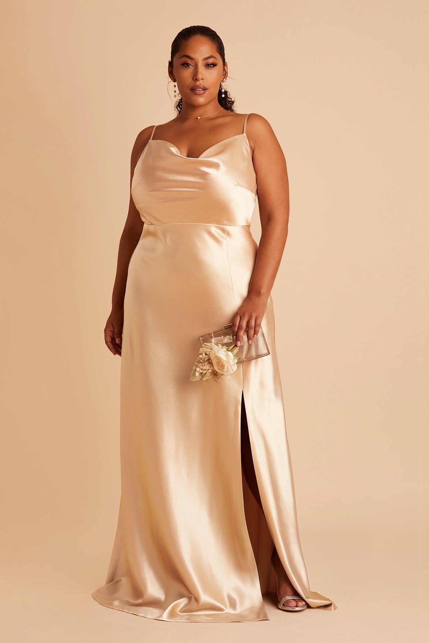 Front view of the Lisa Long Dress Curve in gold satin shows a model revealing their leg and foot in the mid-thigh high slit. They wear the Pearl Pendant Necklace in gold and the Lowell Pearl Drop Earrings. 
