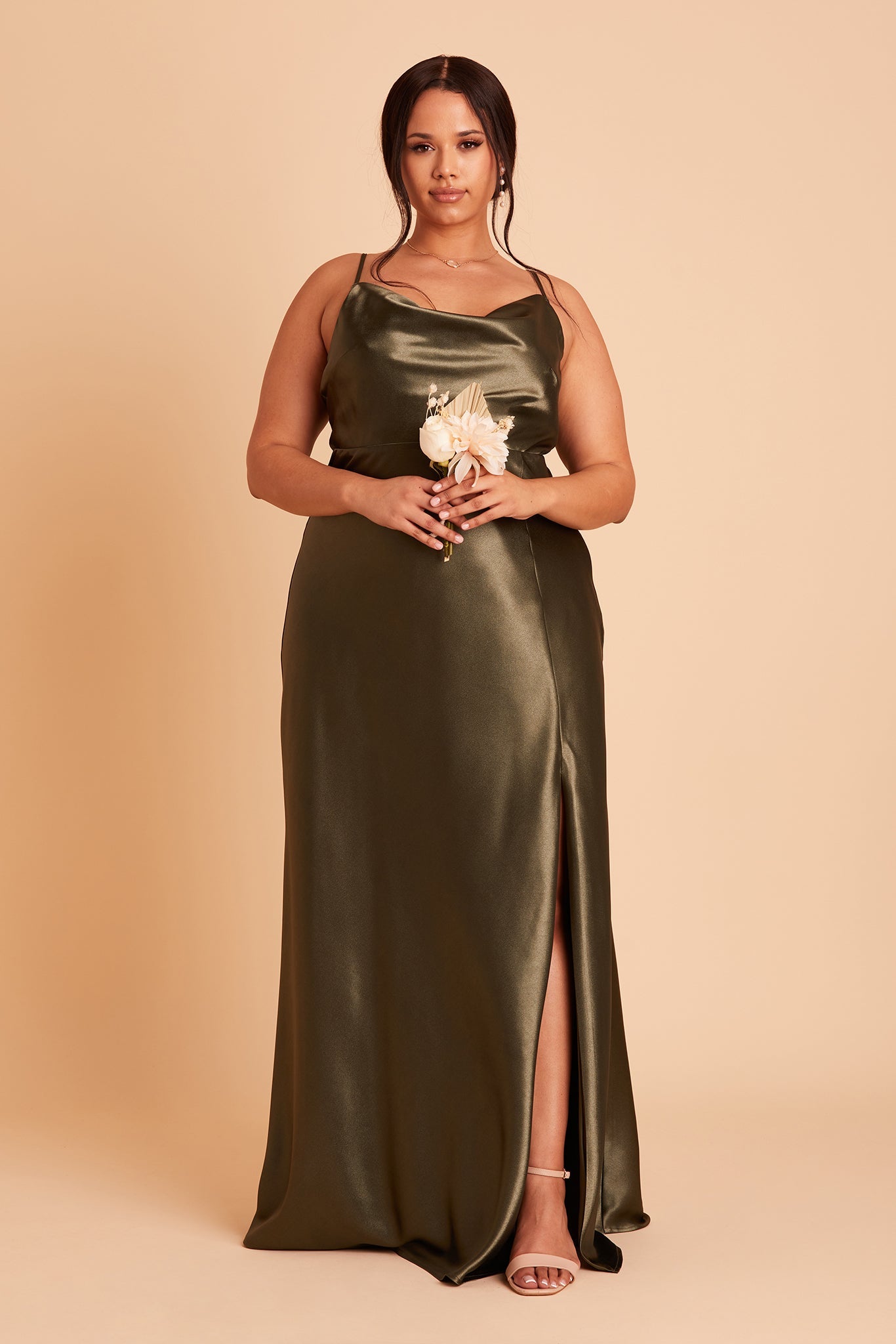 Front view of the Lisa Long Dress Curve in olive satin with a slit shows a full-figured model with a medium skin tone wearing a lightly draped cowl neck bodice with spaghetti straps and a floor length flared dress with a slit. 