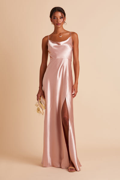 Front view of the Lisa Long Dress in rose gold satin with a slit shows a slender model with a medium skin tone wearing a lightly draped cowl neck bodice with spaghetti straps and a floor length flared dress with a slit. 