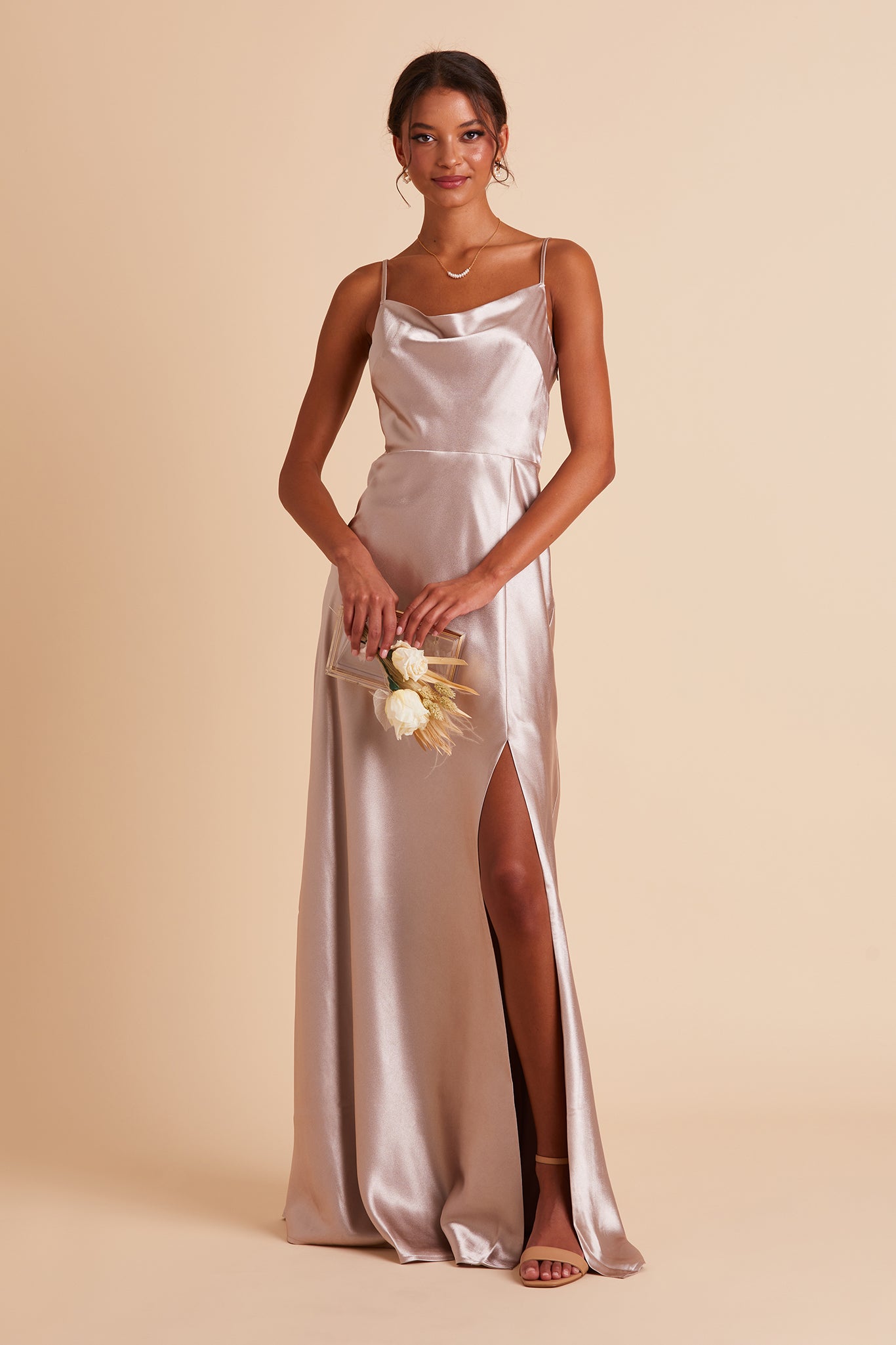 Front view of the Lisa Long Dress in taupe satin shows a model revealing their leg and foot in the mid-thigh high slit. They wear the Natalie Chunky Heel shoe in rose mauve.