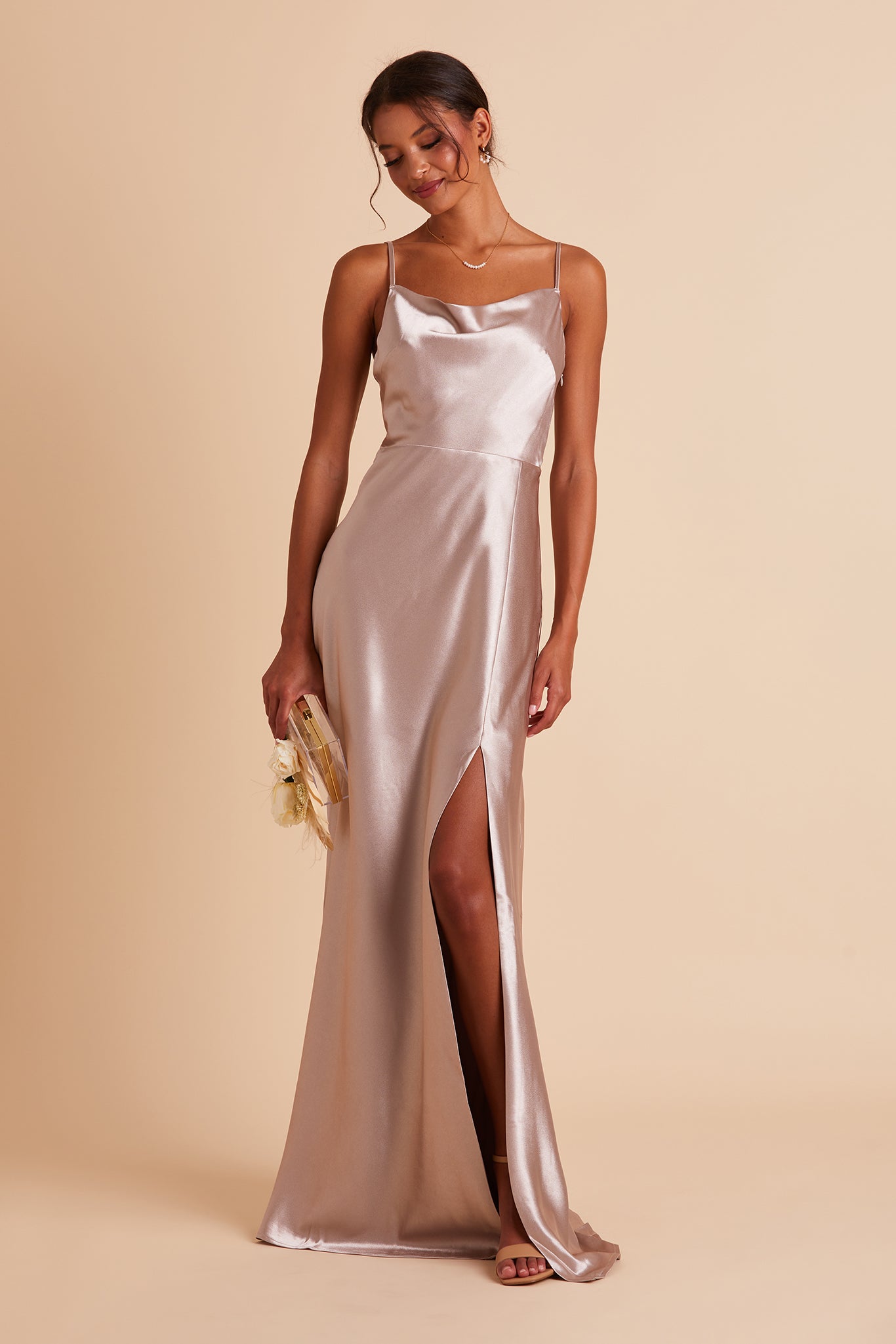 Front view of the Lisa Long Dress in taupe satin shows a slender model with a medium skin tone wearing a lightly draped cowl neck bodice with spaghetti straps and a floor length flared dress with a slit. 