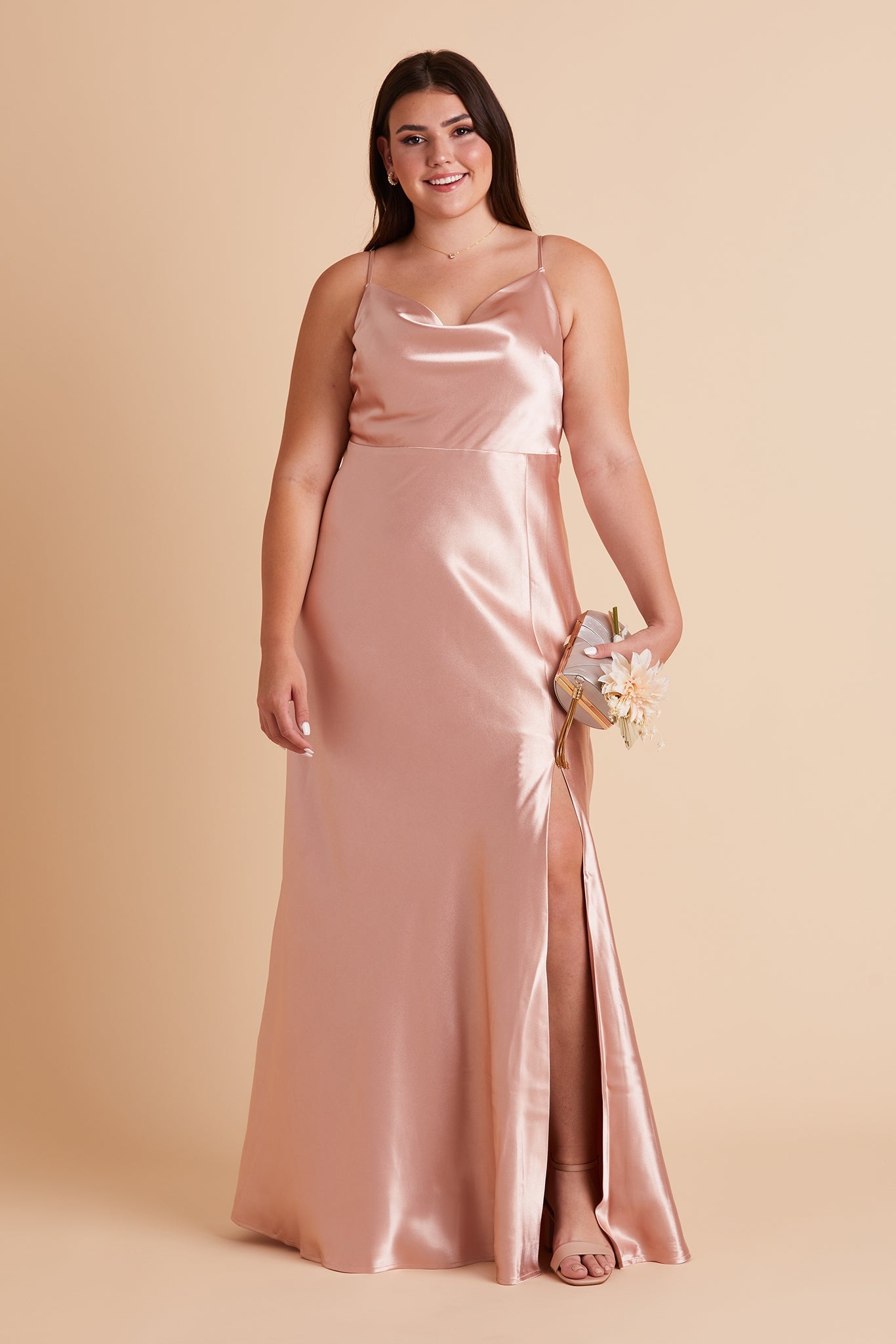 Front view of the Lisa Long Dress Curve Plus Size in rose gold satin shows a model revealing their leg and foot in a strappy heel in the mid-thigh dress slit. 