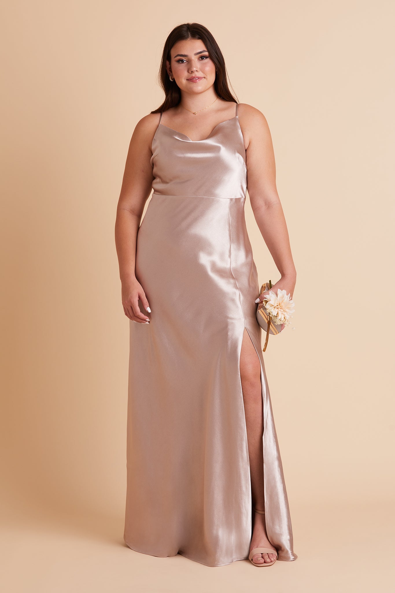 Front view of the Lisa Long Dress Curve in taupe satin shows a full-figured model with a light skin tone wearing a lightly draped cowl neck bodice with spaghetti straps and a floor length flared dress with a slit. 