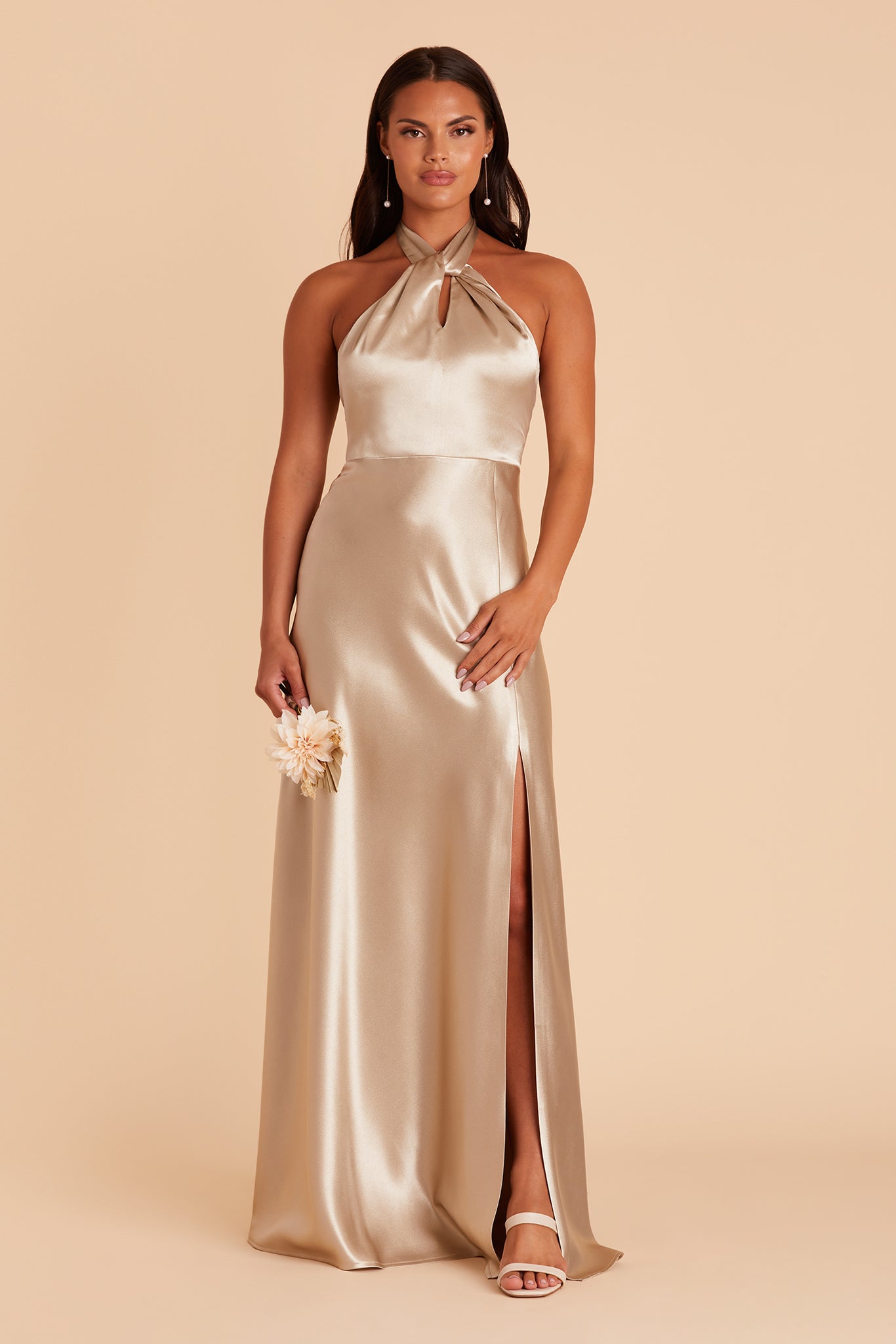 Front view of the Monica Dress in neutral champagne satin shows the model revealing their leg and foot through the dress slit. The model wears the Low Alby Heel shoe.