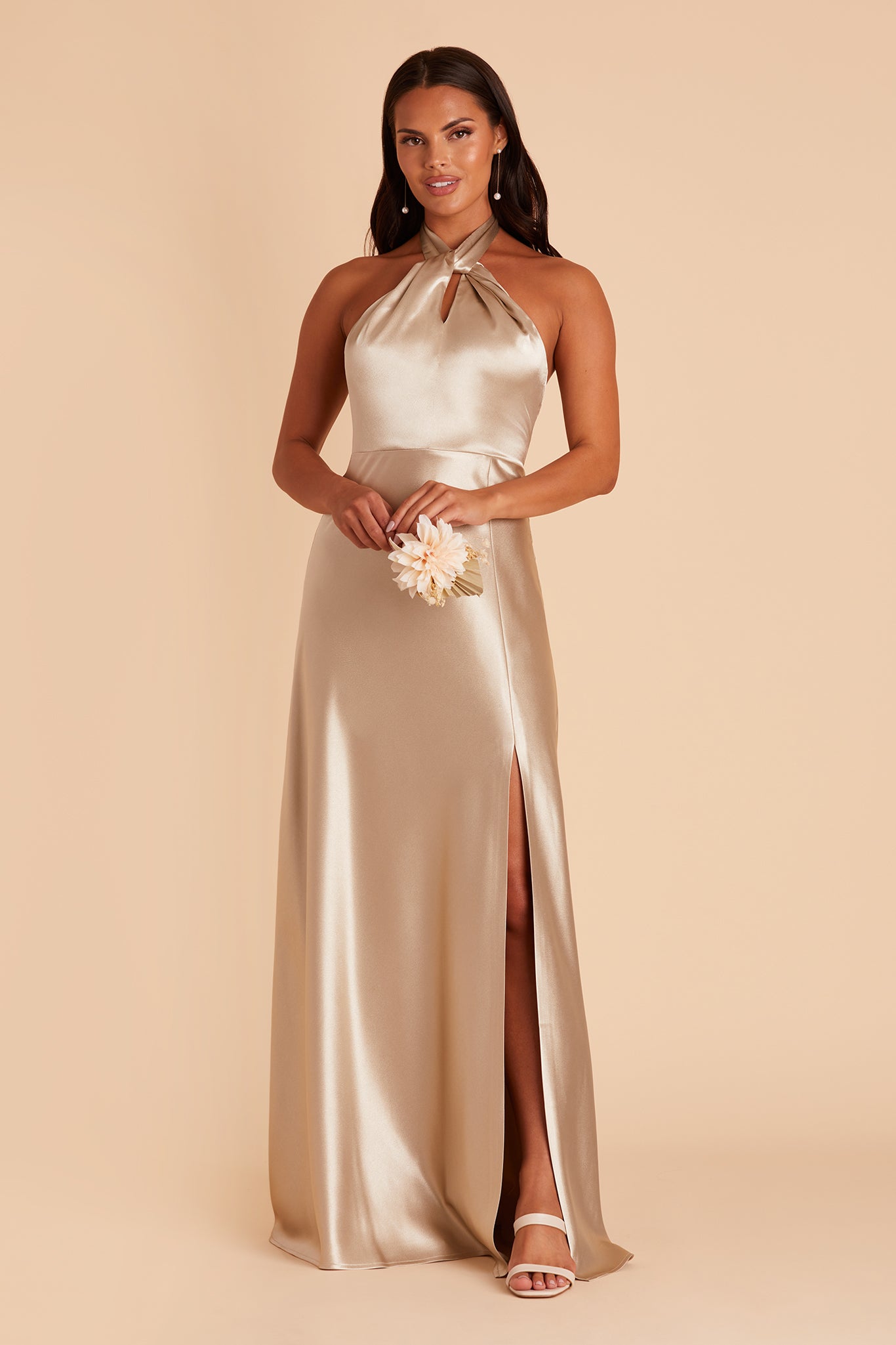 Front view of the Monica Dress in neutral champagne satin shows a model with a medium skin tone wearing an A-line floor length dress with a left slit. The Grecian twist front halter with tie back creates a dress bodice that conforms to the bosom and waist with a smooth fit at the dress skirt top. 