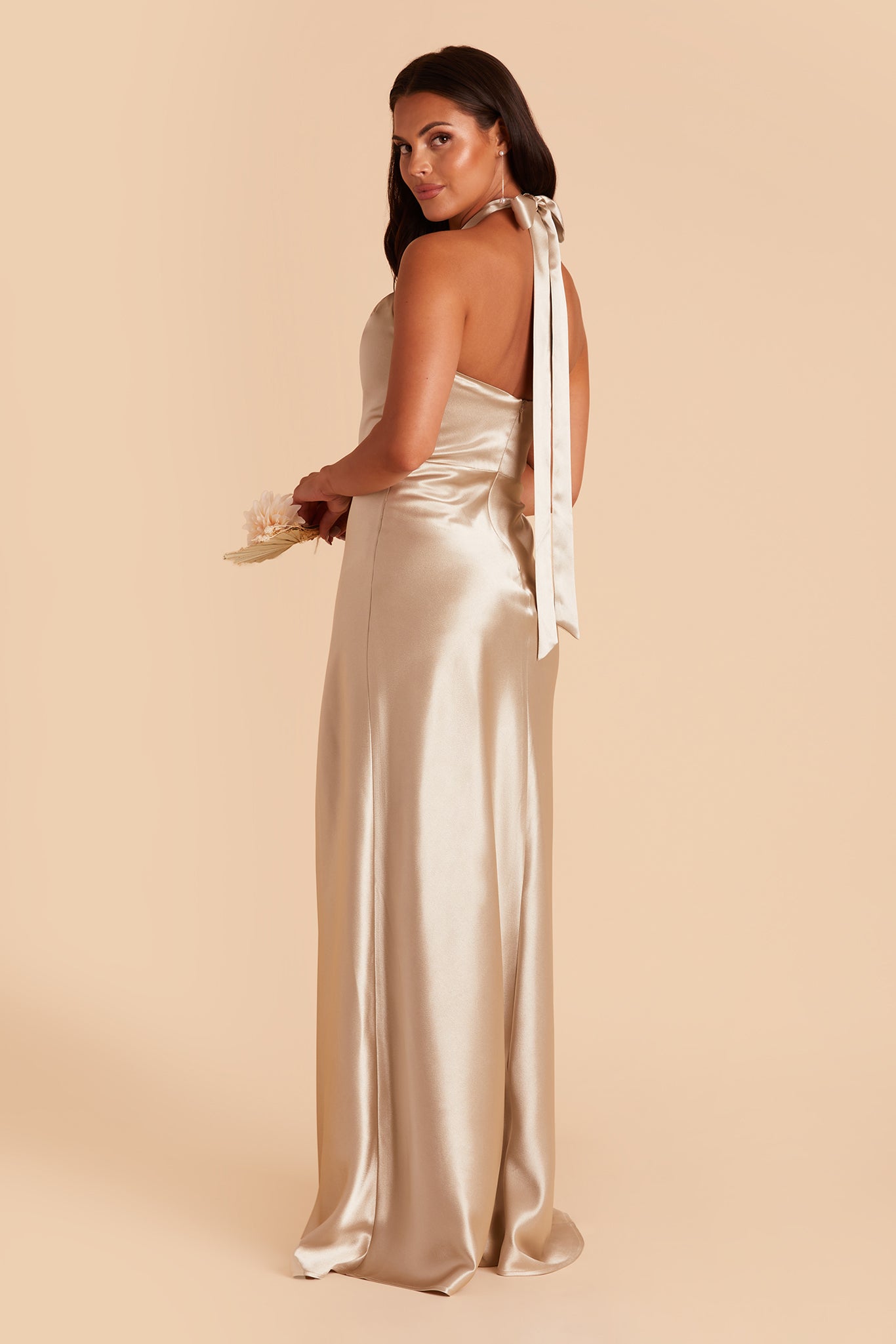 Side view of the Monica Dress in neutral champagne satin shows a model with the halter back tied in a bow at the back of their neck with the long tie ends draping along the back to the backside. The dress conforms to the model's body along the back and hips flaring slightly to the feet.