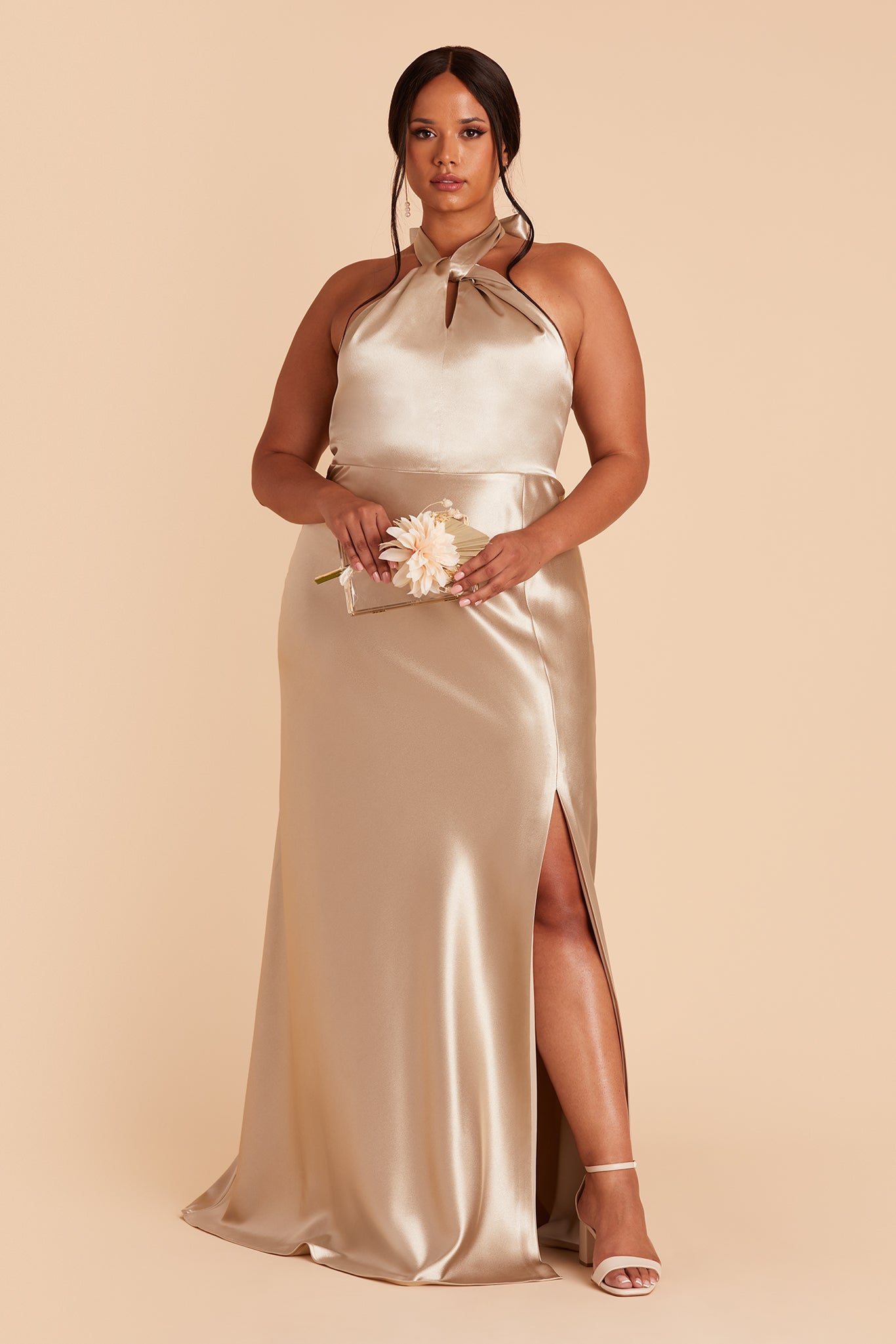 Front view of the Monica Dress Curve in neutral champagne satin shows a full-figured model with a medium skin tone wearing an A-line floor length dress with a left slit. The Grecian twist front halter with tie back conforms to the bosom and waist with a smooth fit at the dress skirt top. 