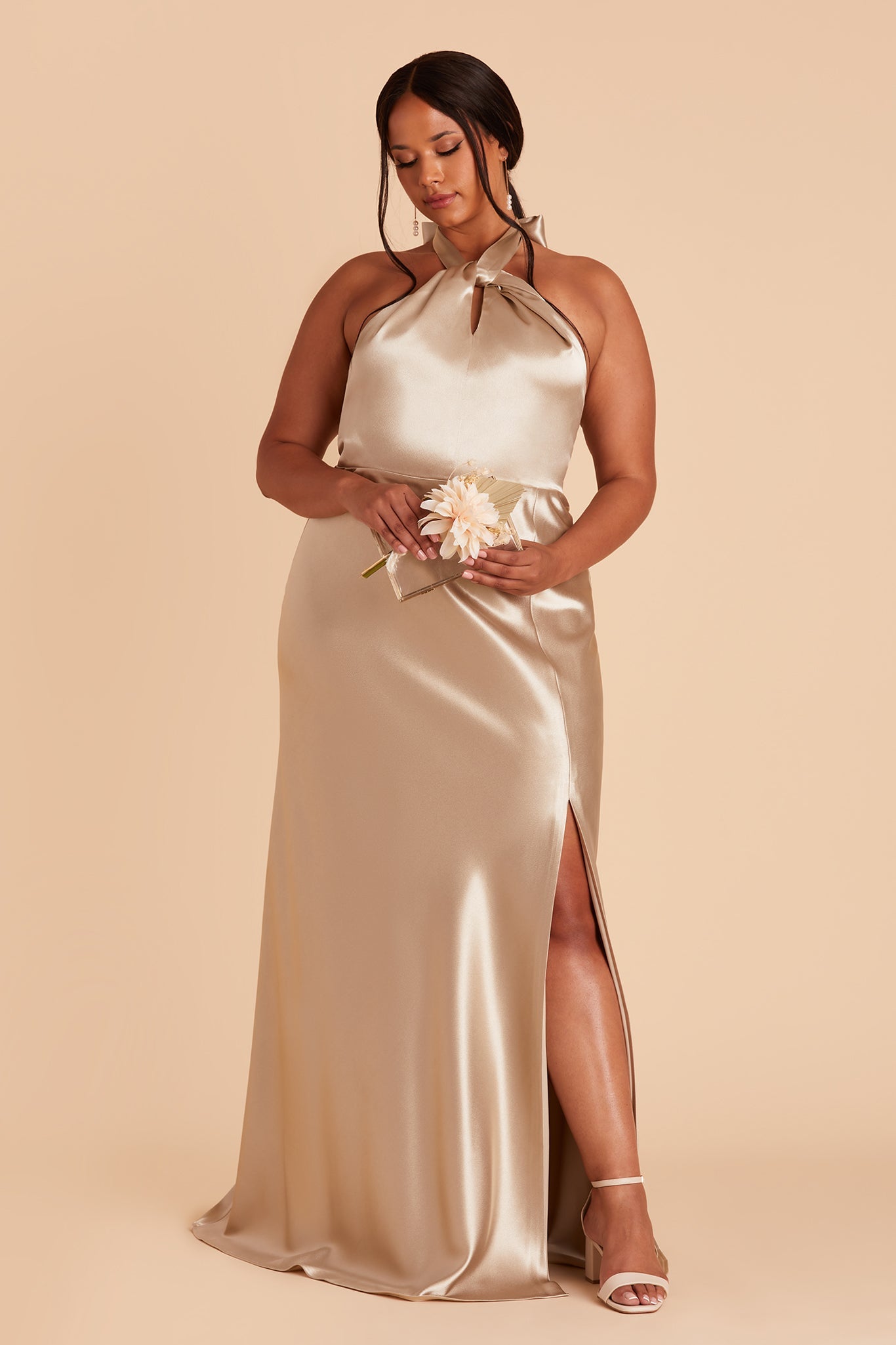 Front view of the Monica Dress Curve in neutral champagne satin shows the model revealing their leg and foot through the dress slit. The model wears the Natalie Chunky Heel in almond.
