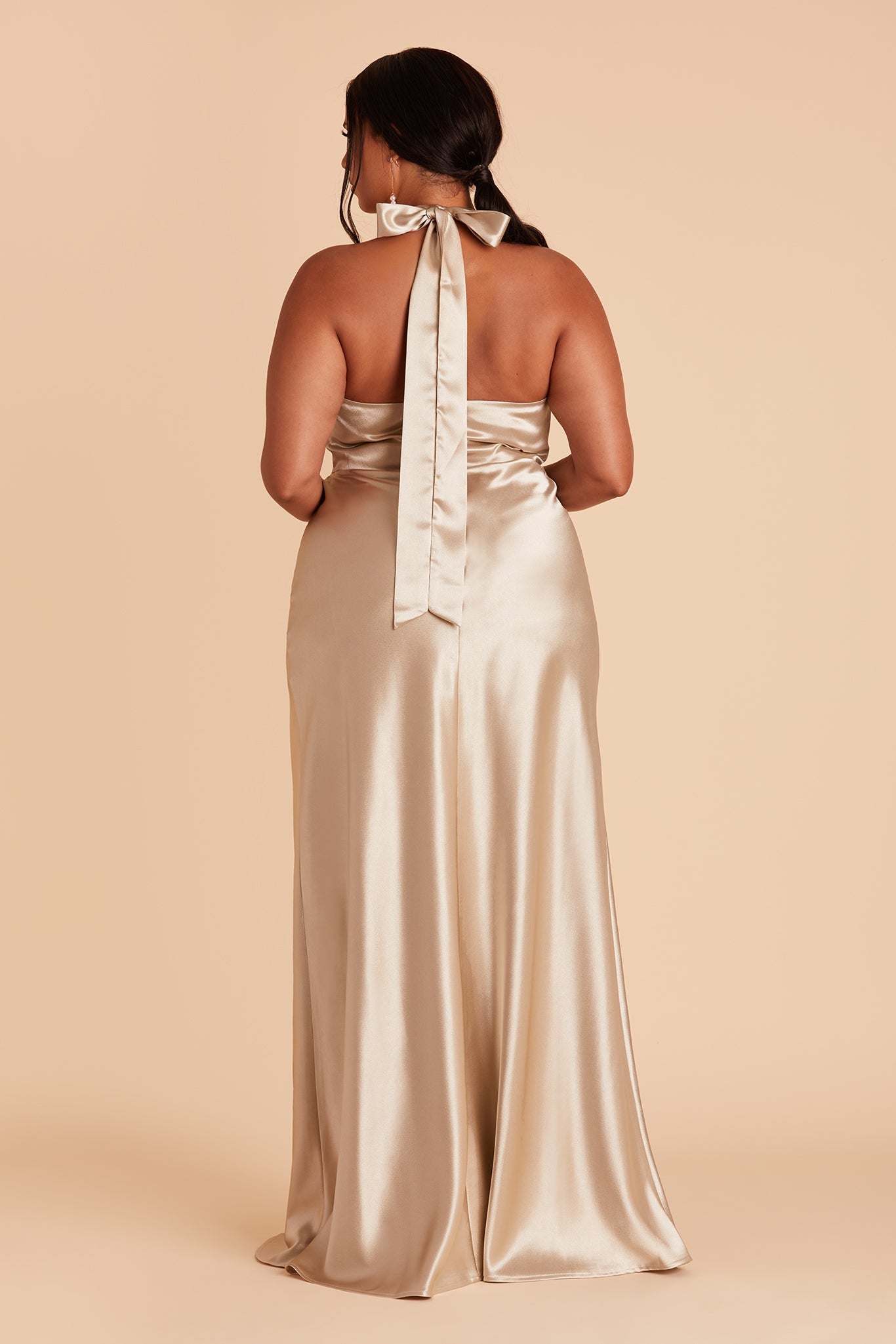 Back view of the Monica Dress Curve in neutral champagne satin shows a model with a halter back tied in a bow at the back of their neck with the long tie ends draping along the back to the backside. The dress conforms to their body along the back and hips flaring slightly to the feet.