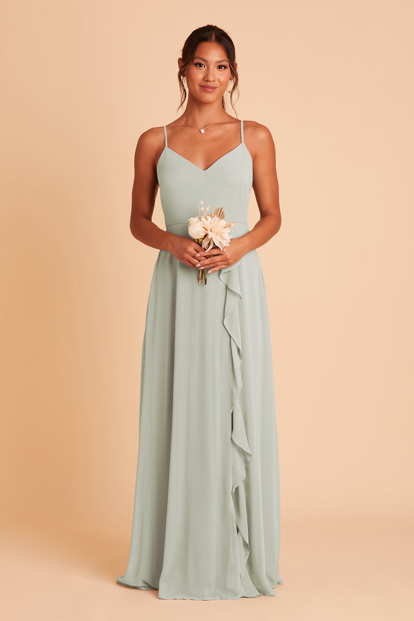 Front view of the Theresa Dress in sage chiffon with the optional slit shows a slender model with a medium skin tone wearing a floor length, A-line dress with a sweeping column skirt and ruffle along the slit of the skirt. The fitted V-neck bodice with spaghetti straps. 