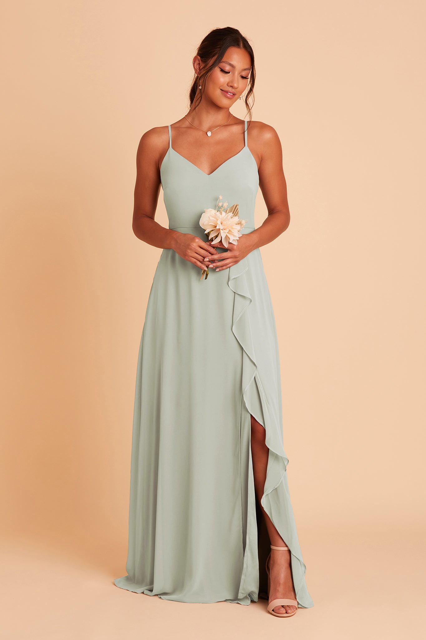 Front view of the Theresa Dress in sage chiffon with the optional slit shows a model holding a dried flower bouquet in ivory at waist height above the sweeping column skirt and slit ruffle. The model reveals her lower leg through the slit and wears the Mary High Chunky Heel shoe. 