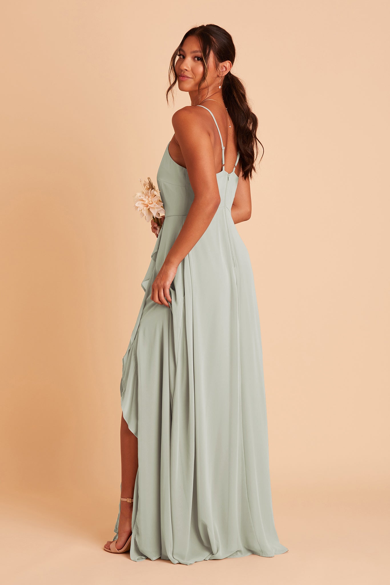 Side view of the Theresa Dress in sage chiffon with the optional slit shows a model looking at us and holding the side slit to reveal the ruffles and high heel shoes in nude blush. The adjustable shoulder straps are angled toward the center seam.