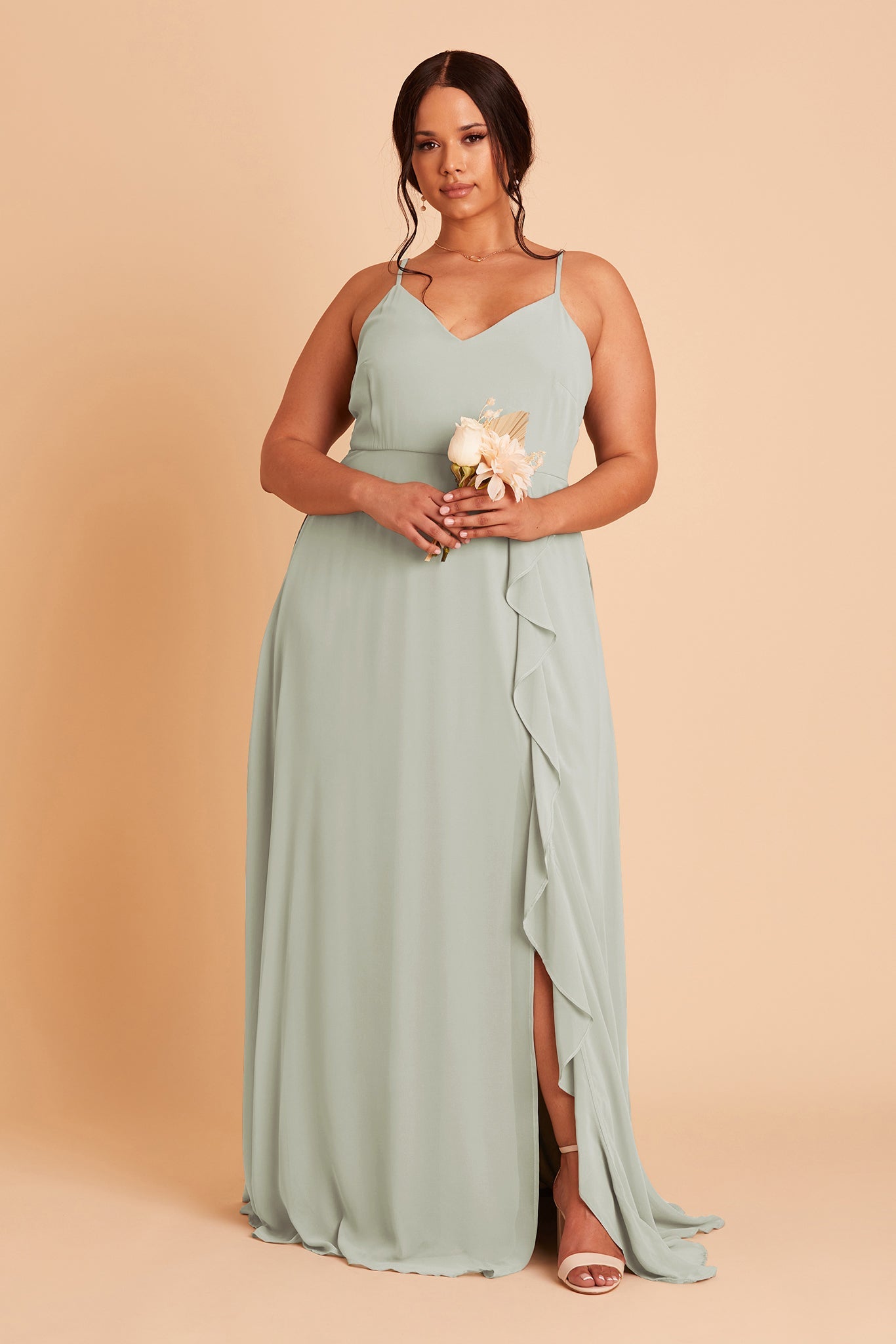 Front view of the Theresa Dress Curve in sage chiffon with the optional slit shows a full-figured model with a medium skin tone wearing a floor length, A-line dress with a sweeping column skirt and ruffles along the slit of the skirt. The fitted V-neck bodice with spaghetti straps. 