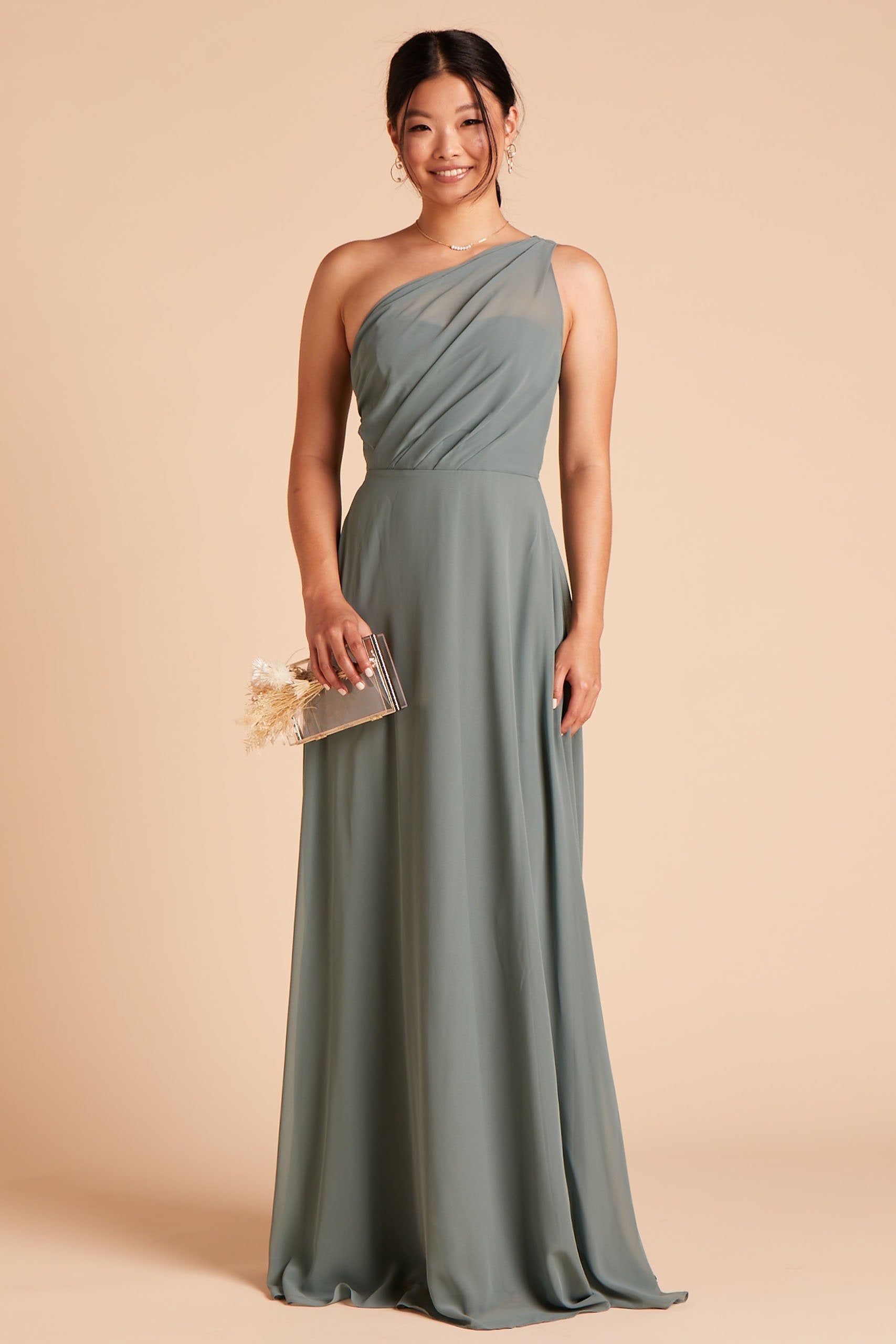 Front view of the Kira Dress in sea glass chiffon without the optional slit shows a slender model with a medium skin tone wearing an asymmetrical one-shoulder, full-length dress. Soft pleating gathers at the left shoulder of the bodice with a smooth fit at the waist as the dress skirt with a slight A-line silhouette flows to the floor.