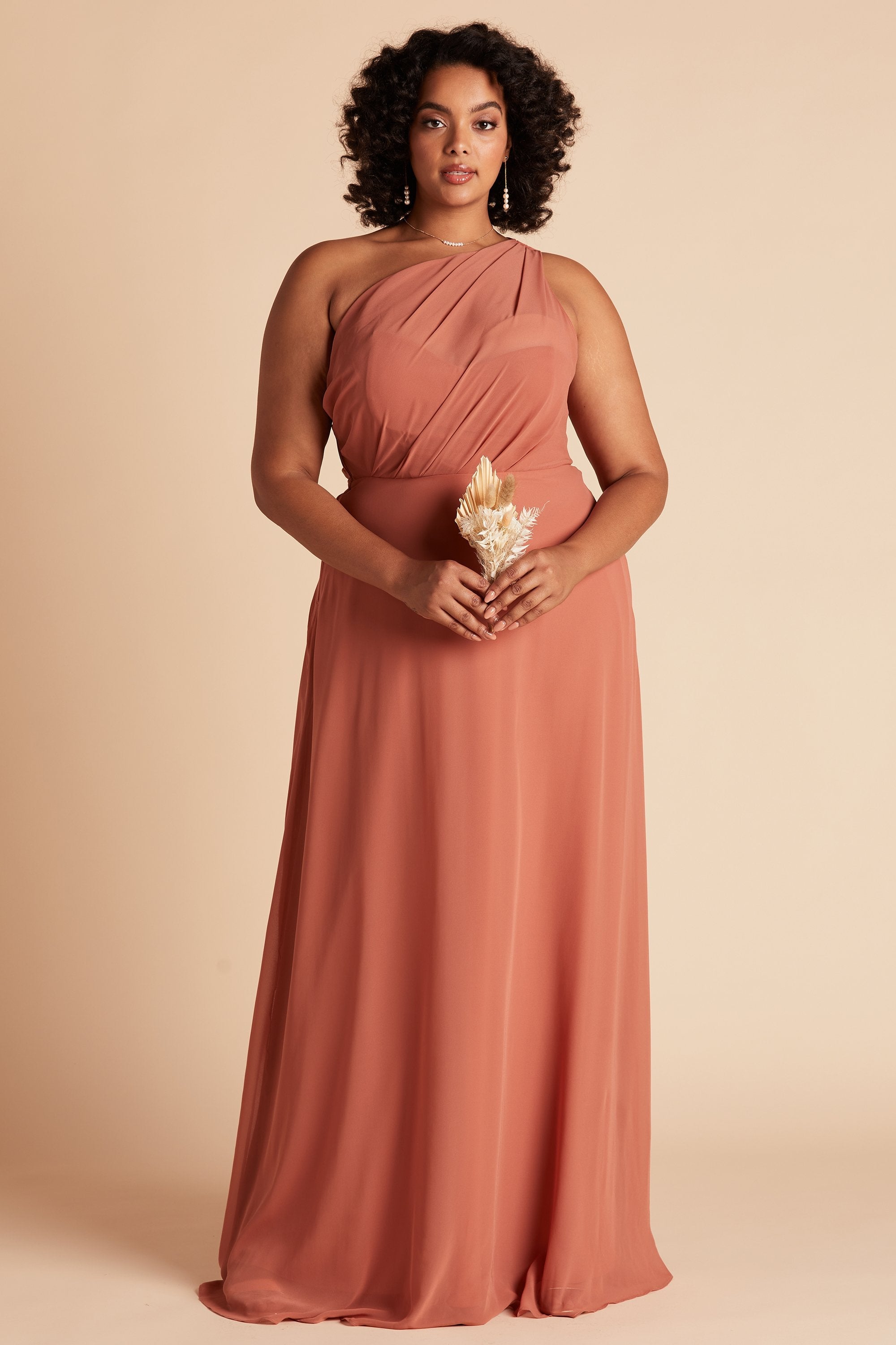 Front view of the Kira Dress Curve in terracotta chiffon without the optional slit shows a full-figured model with a medium skin tone wearing an asymmetrical one-shoulder, full-length dress. Soft pleating gathers at the left shoulder of the bodice with a smooth fit at the waist as the dress with a slight A-line silhouette flows to the floor.