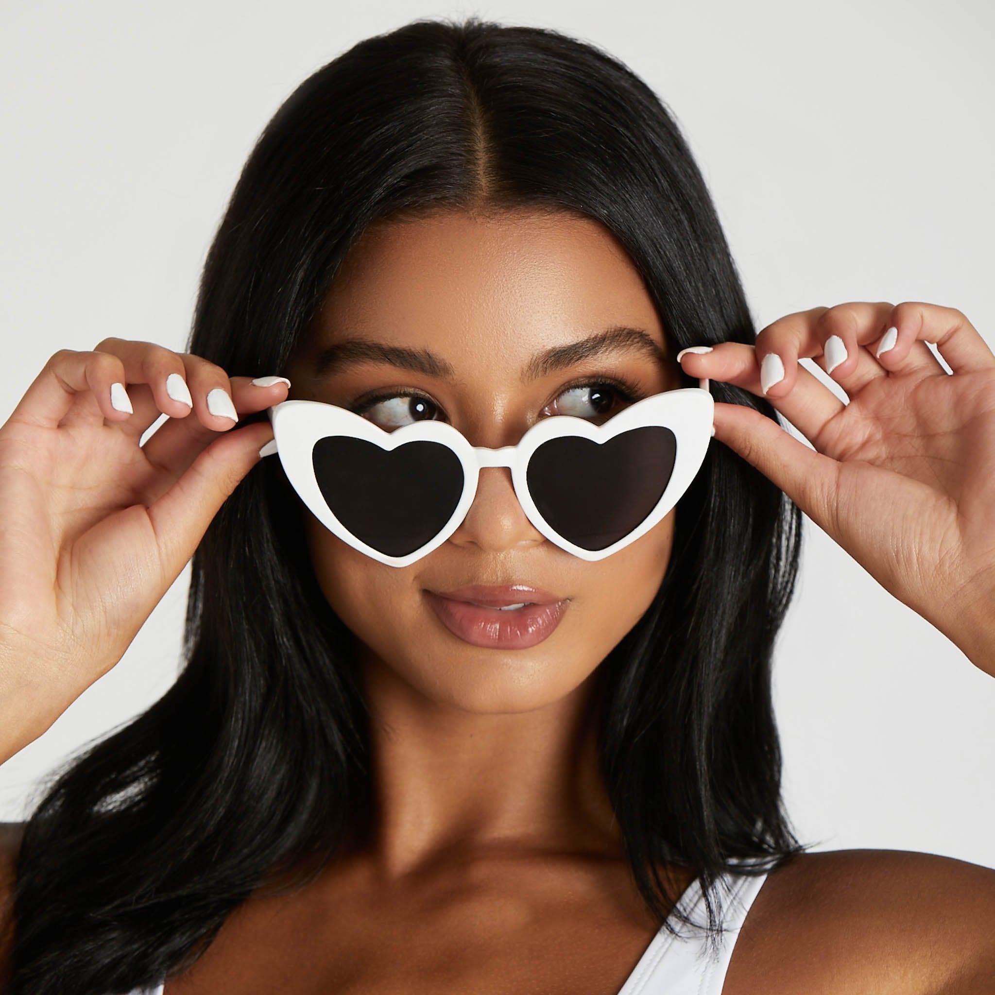 Front view of a model with a medium skin tone wearing Heart Sunglasses in white. She lowers the lenses to look over the top of the frames.