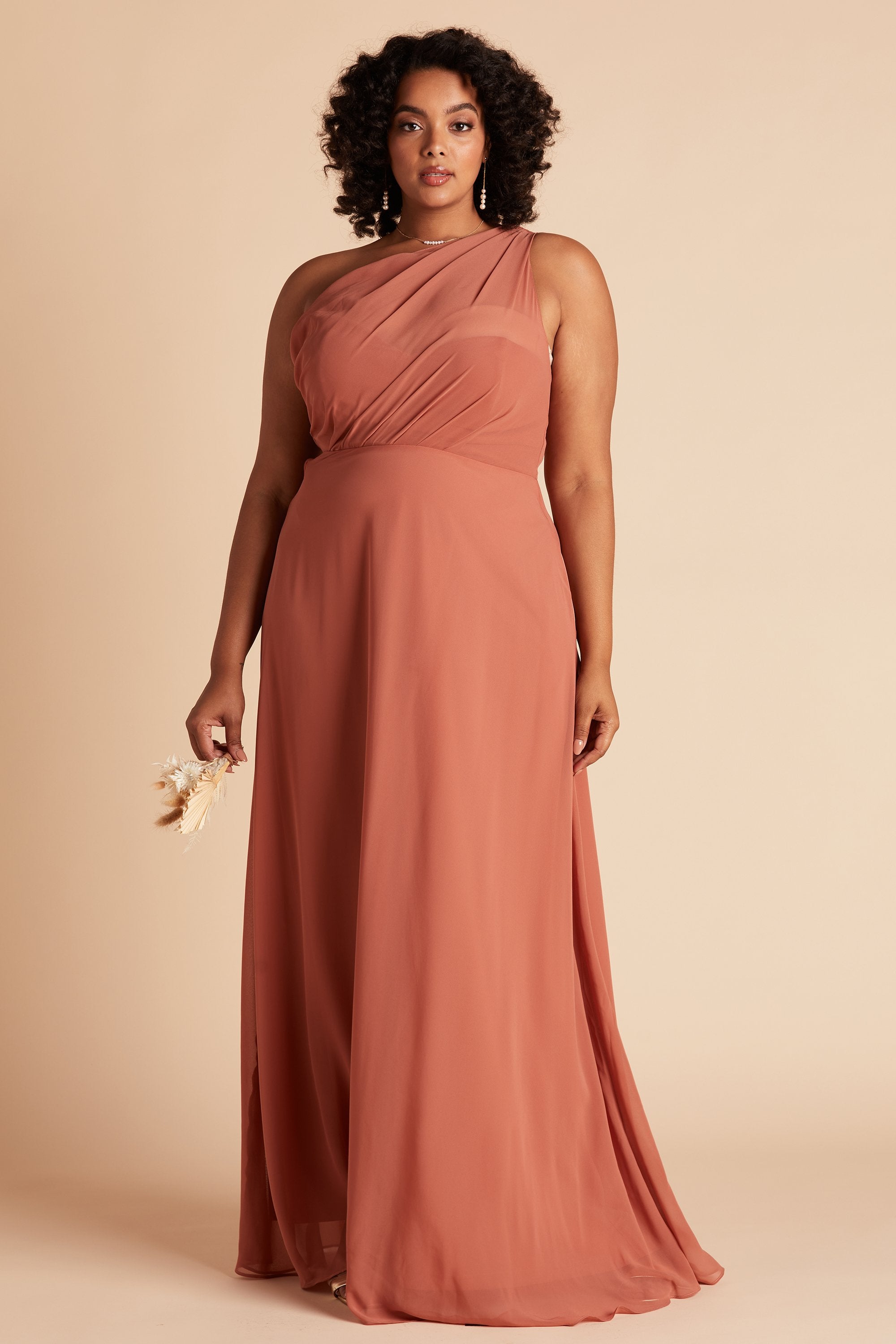 Front view of the Kira Dress Curve in terracotta chiffon without the optional slit shows a full-figured model with a medium skin tone. The asymmetrical, chiffon one-shoulder bodice wraps across their full bosom. Soft pleating gathers at the left shoulder of the bodice with a smooth fit at the waist as the dress skirt flows to the floor.