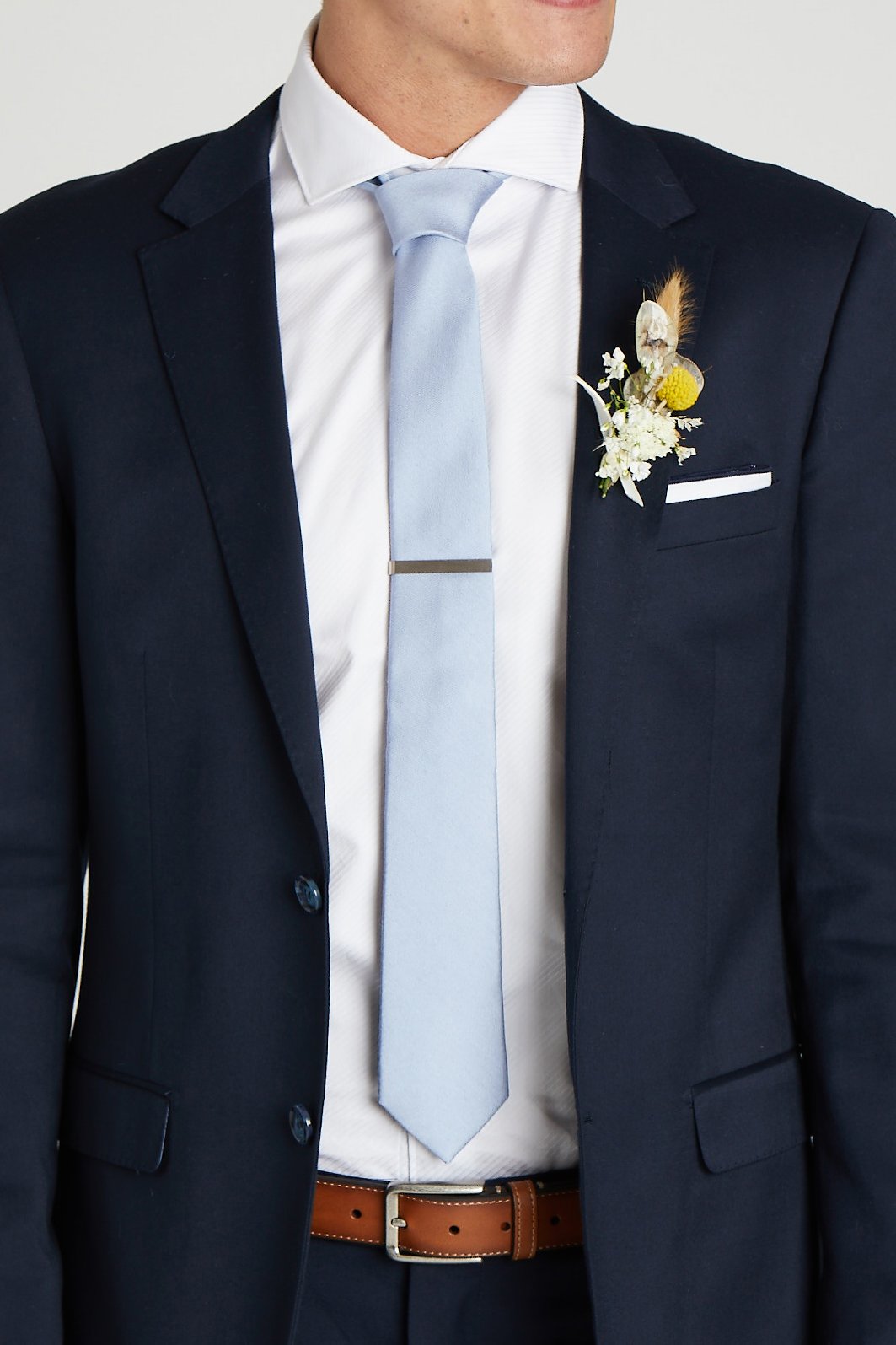 Front closeup view of the model wearing the Simon Necktie in dusty blue with a white button down collared shirt and navy blue suit. The tie is kept in place with a narrow rectangular tie clasp in matte silver.