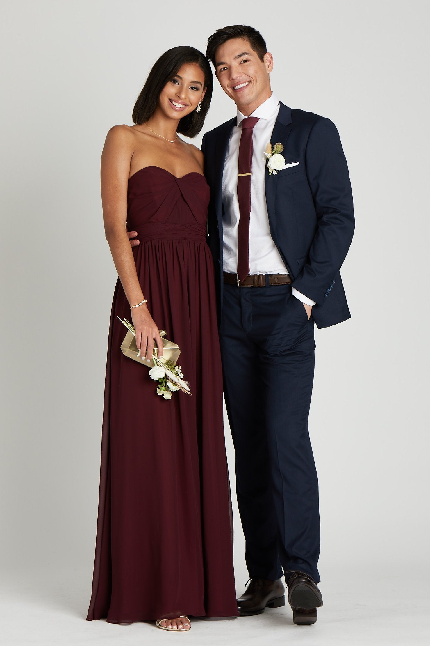 Front view of two models in a side embrace at the waist. One model with a slender physique and a medium skin tone wears the Simon Necktie in cabernet with a white collared button down shirt and the Navy Blue Suit by Suitshop. The second model with a slender figure and a medium skin tone coordinates their look wearing the floor length Spence Convertible Dress in cabernet.