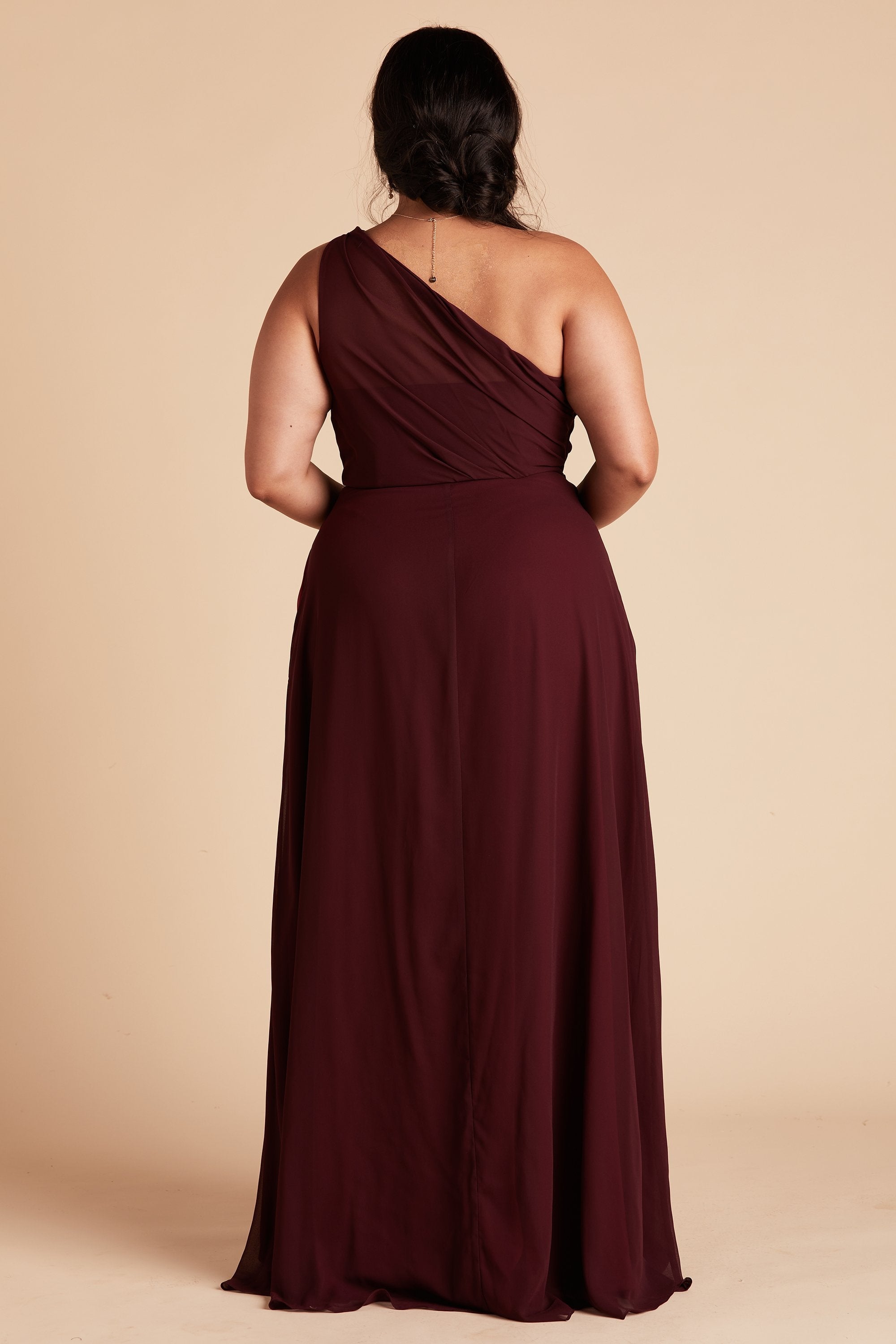 Back view of the Kira Dress Curve in cabernet chiffon shows a full-figured model with a medium skin tone. The back of the dress has sheer chiffon fabric which creates a pleated Grecian one-shoulder neckline and bodice. 