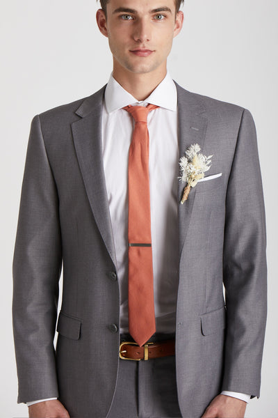 Front closeup view of a model wearing the Simon Necktie in terracotta with a white button down collared shirt and medium grey suit. The tie is kept in place with a narrow rectangular tie clasp in matte gold.
