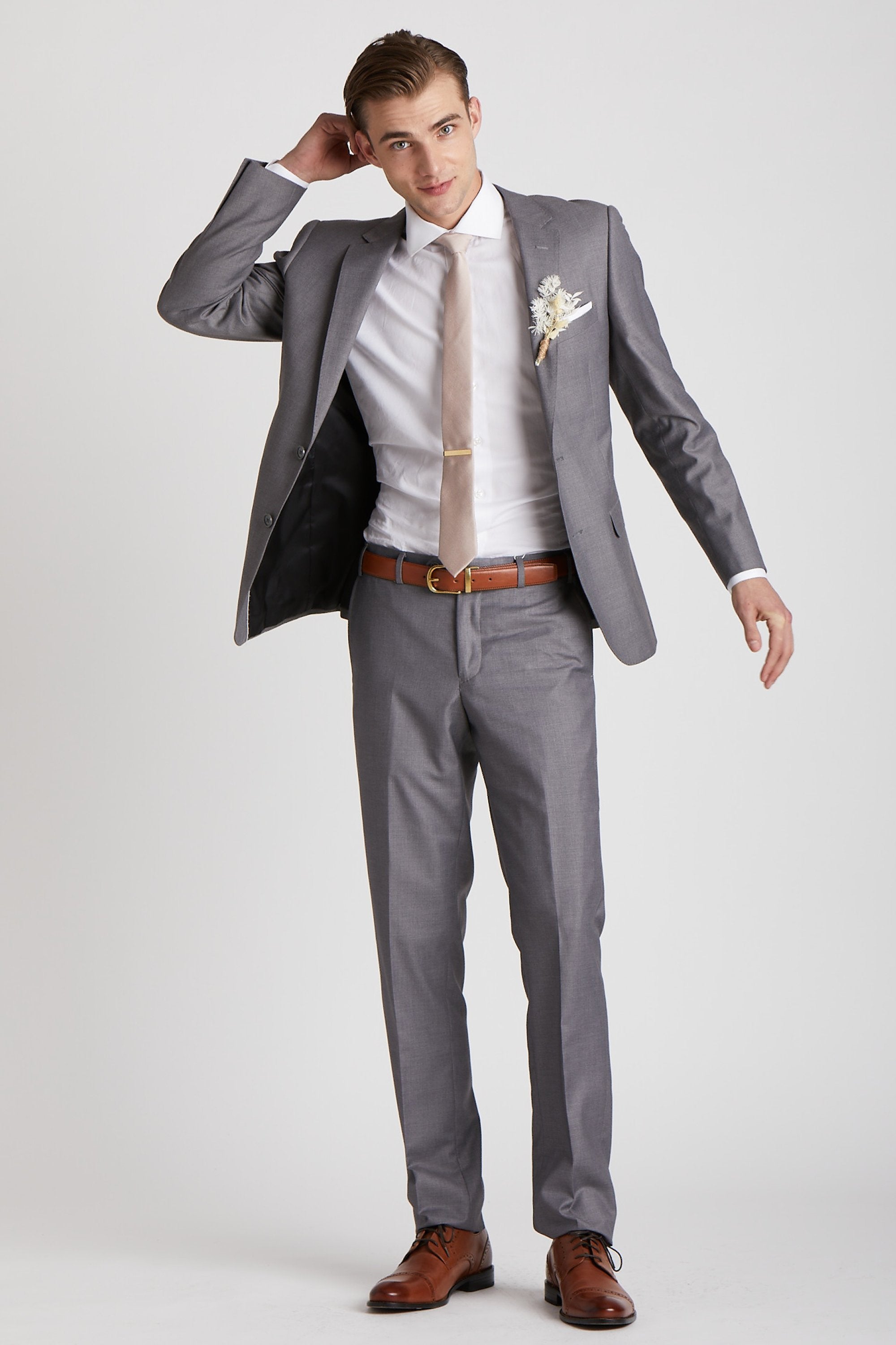 Front full view of a model wearing the Simon Necktie in taupe coordinated with a medium grey suit with a white collared shirt, medium brown shoes and belt, white pocket square, and a dried flower boutonniere in ivory. 