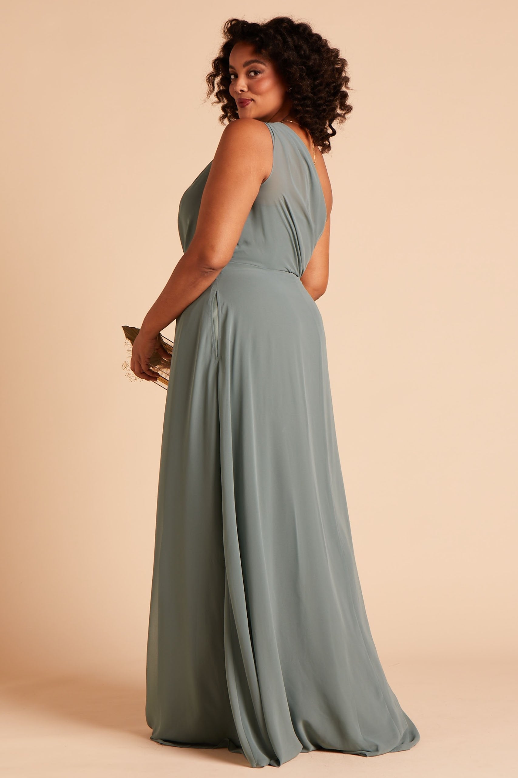 Back and side view of the floor-length Kira Dress Curve in sea glass chiffon shows a full-figured model with medium skin wearing a sheer, softly pleating chiffon gathered at the bodice shoulder and draping across the back to the side seam with an inset pocket.