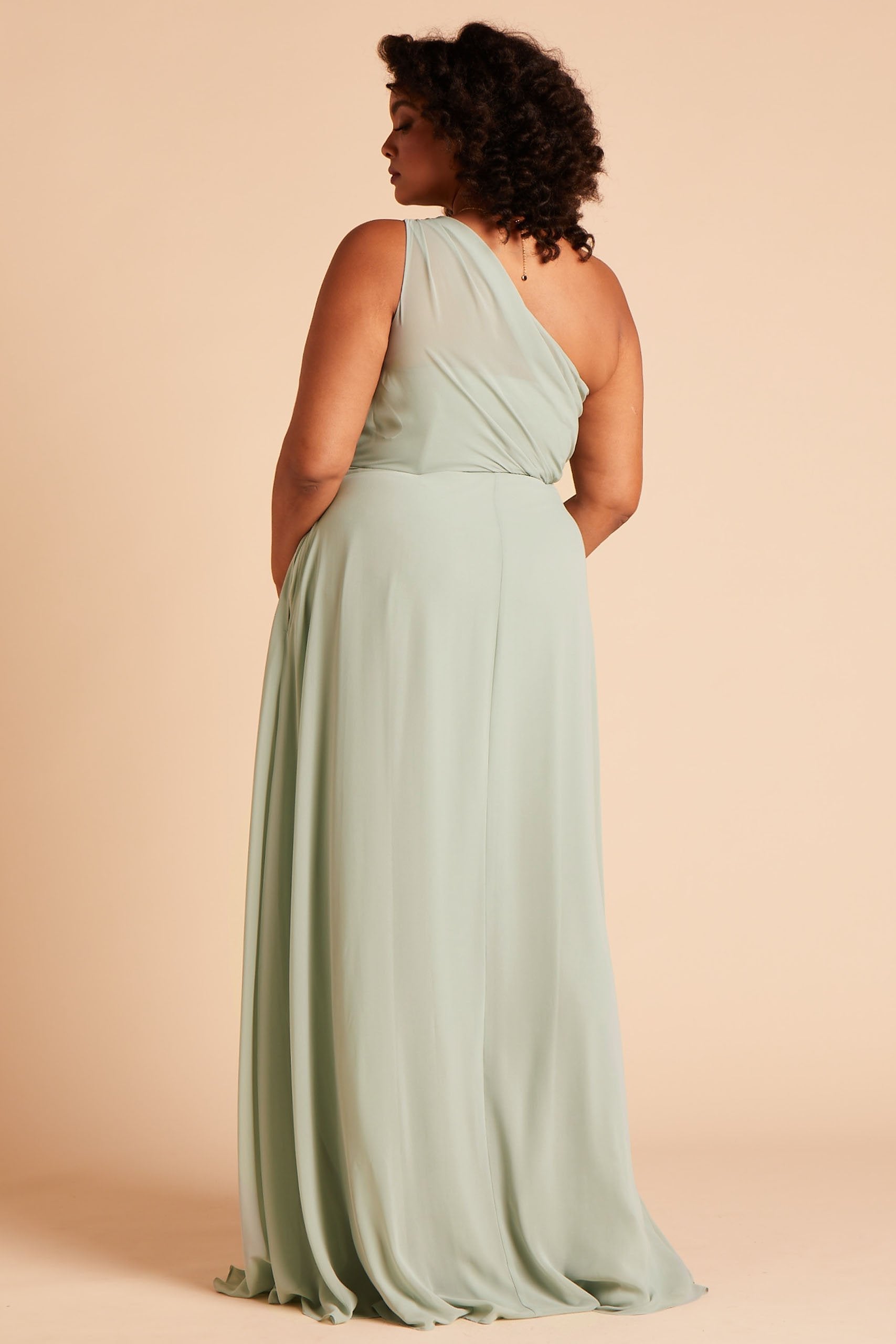 Back and side view of the floor-length Kira Dress Curve in sage chiffon shows a full-figured model with medium skin wearing a sheer, softly pleating chiffon gathered at the bodice shoulder and draping across the back to the side seam with an inset pocket.
