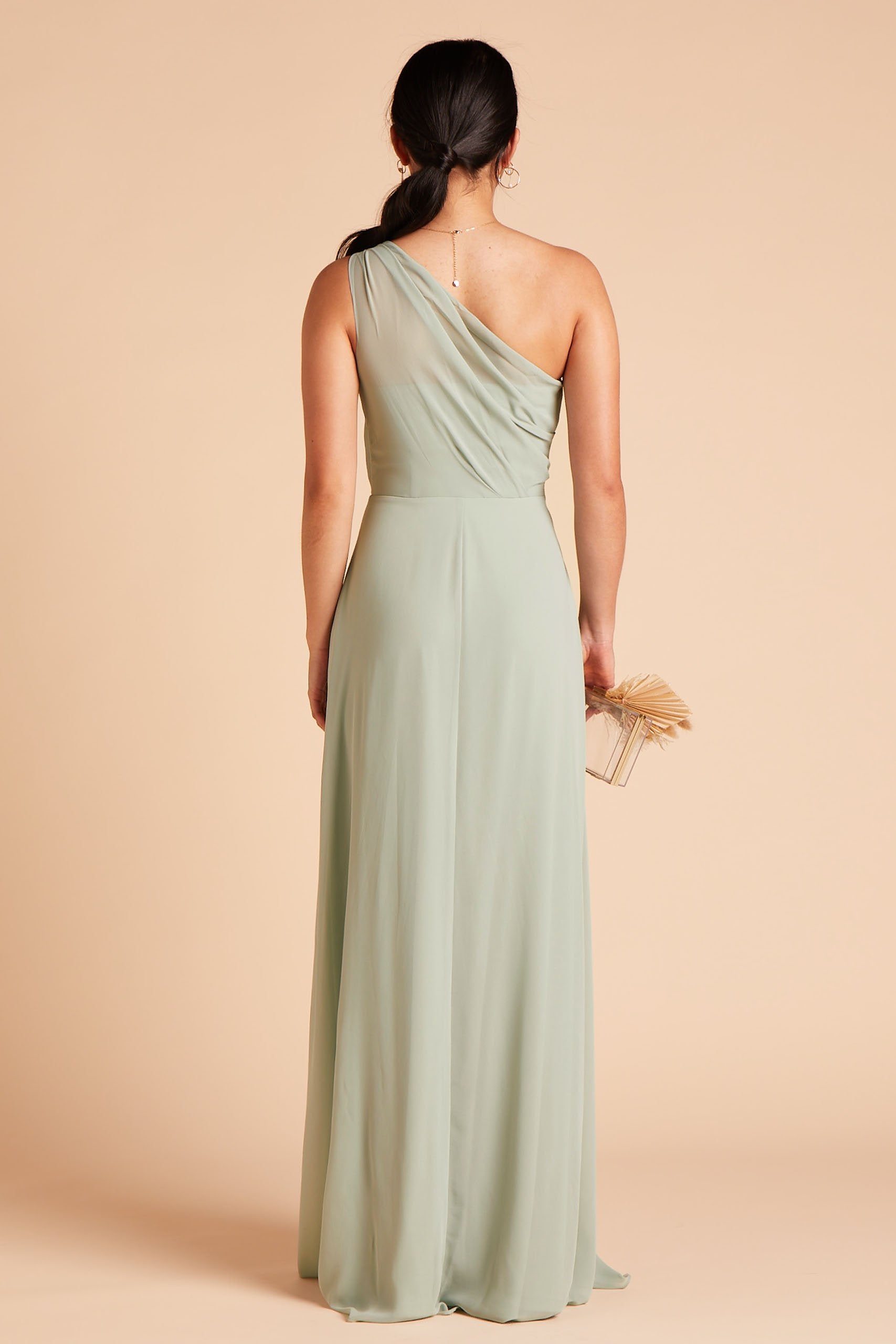 Back view of the Kira Dress in sage chiffon shows a slender model with a medium skin tone. The back of the dress has sheer chiffon fabric which creates a pleated Grecian one-shoulder neckline and bodice.