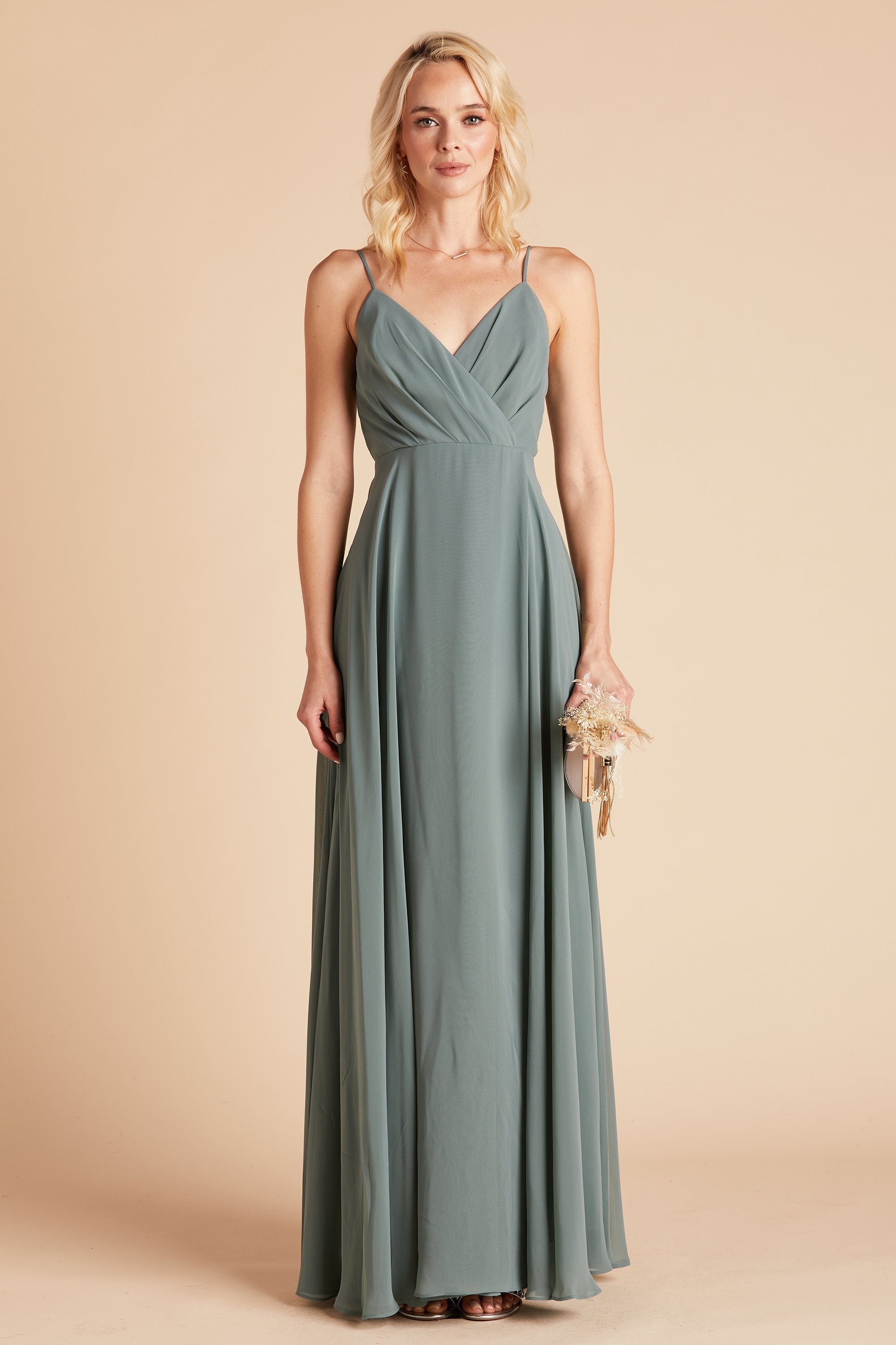 Front view of the Kaia Dress in sea glass chiffon shows the model holding a bouquet and pale pink clutch at their side. The V-neck and wide pleated bodice overlaps across the chest. The skirt front falls smoothly towards the floor, gathering gently at the sides.