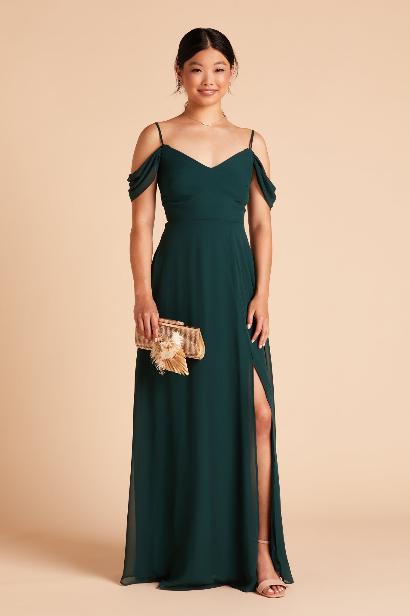 Front view of the floor-length Devin Convertible Bridesmaid Dress in emerald chiffon flaunts the optional thigh-high slit over the left leg as well as the detachable sleeve draping over the upper arms.