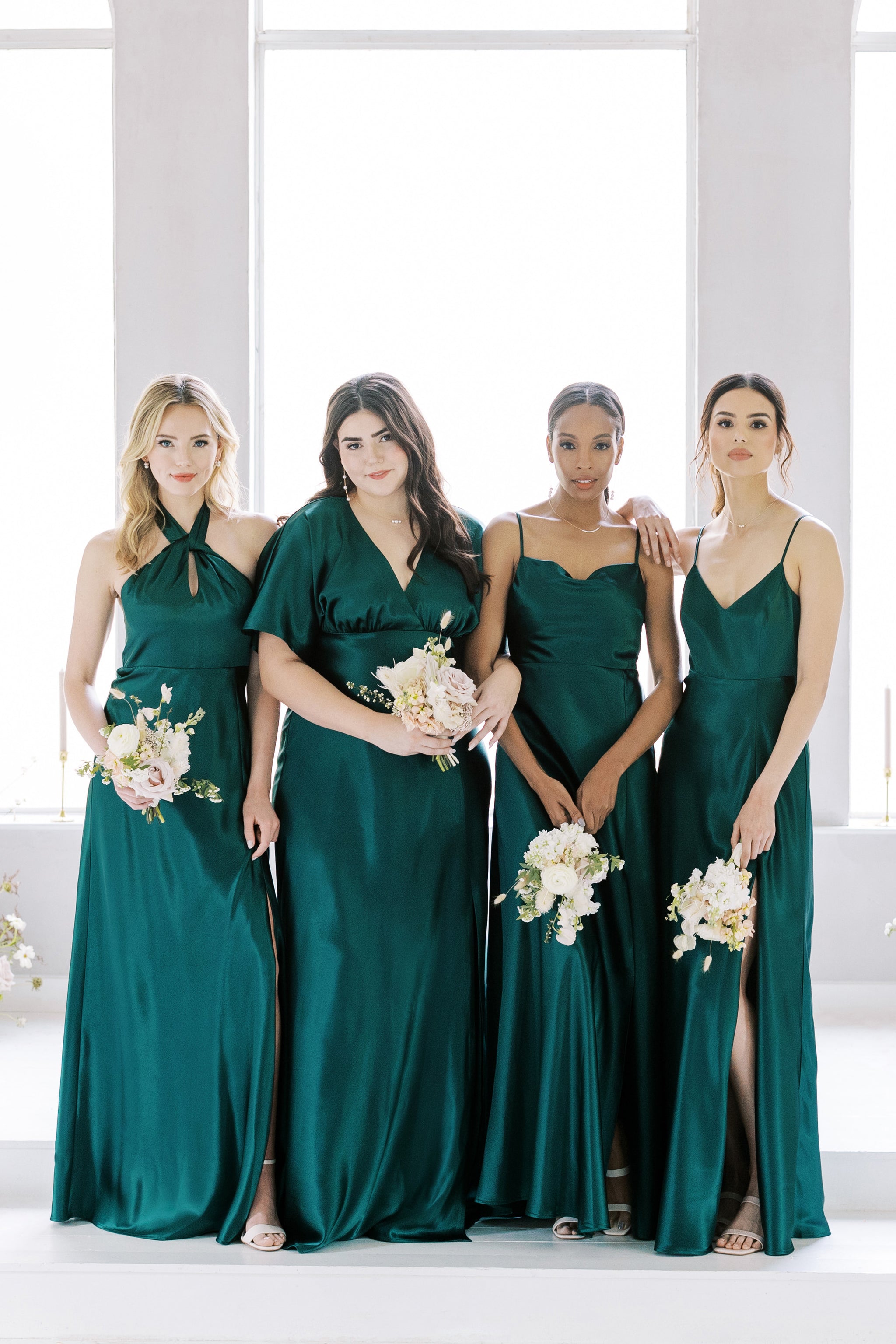 Front view of four models wearing coordinating floor length dresses in emerald satin. The Grace Convertible and the Jay dresses are worn by slender models with a light skin tone. The Lisa Long dress is worn by a slender model with a medium skin tone and the Jesse Dress Curve is worn by a full figured model with a light skin tone. 