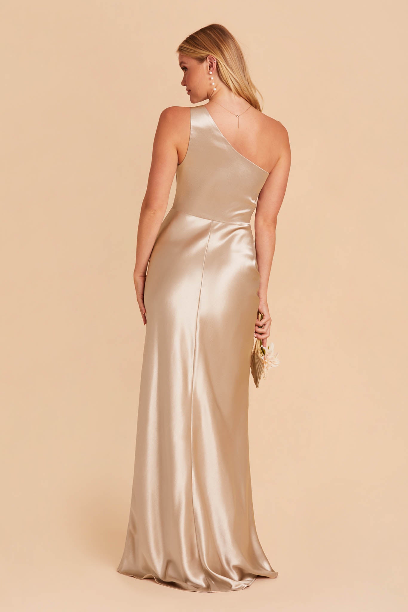 Back view of the Kira Dress in neutral champagne satin shows a slender figured model with a light skin tone. The back of the dress has a smooth fit in the one-shoulder bodice and waist flaring into a mermaid style skirt.  