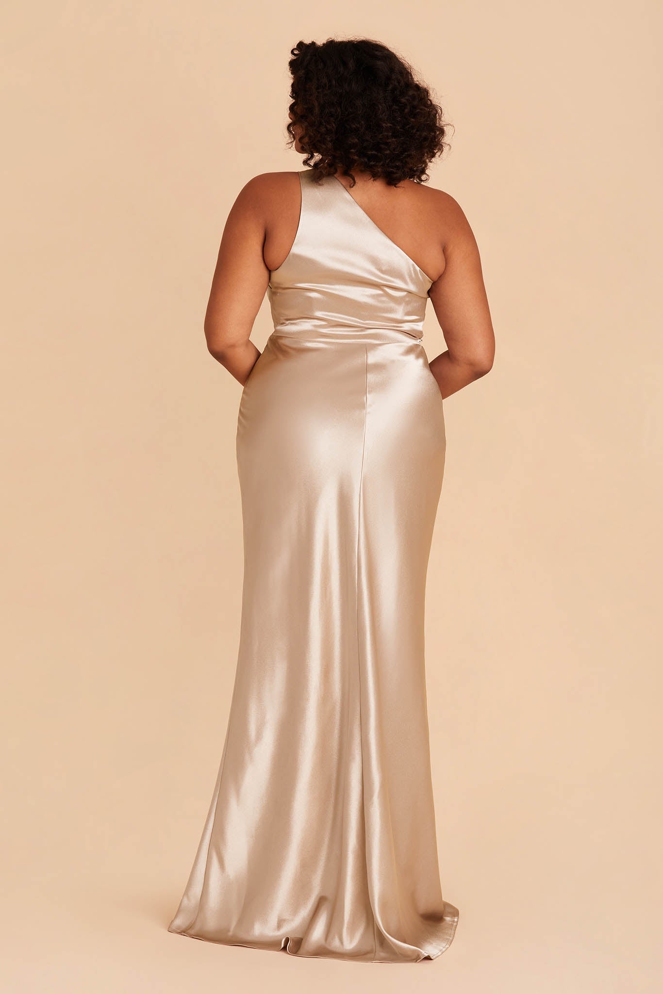 Back view of the Kira Dress Curve in neutral champagne satin shows a full-figured model with a medium skin tone. The back of the dress has a smooth fit across the one-shoulder bodice and waist that attaches to a mermaid-style skirt.  