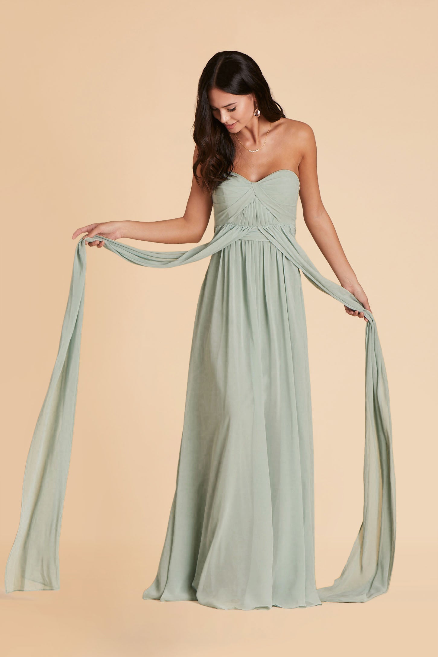 Front view of the Grace Convertible Bridesmaid Dress in sage chiffon as the model holds a front streamer connected at the waist in each hand. The streamers are extra long, longer than the floor-length skirt.
