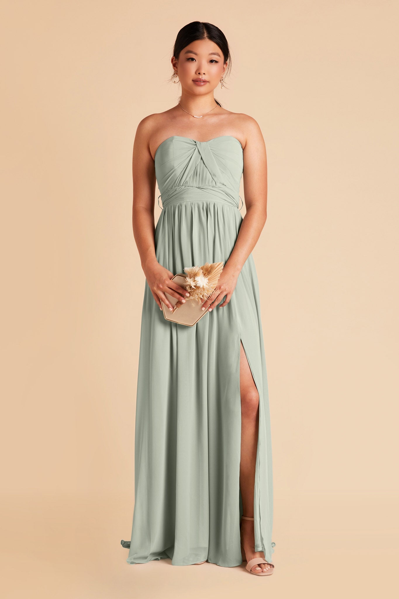 Front view of the Grace Convertible Bridesmaid Dress in sage chiffon on a slender model with light skin whose left leg peeks out through a slit in the flowing floor-length skirt.